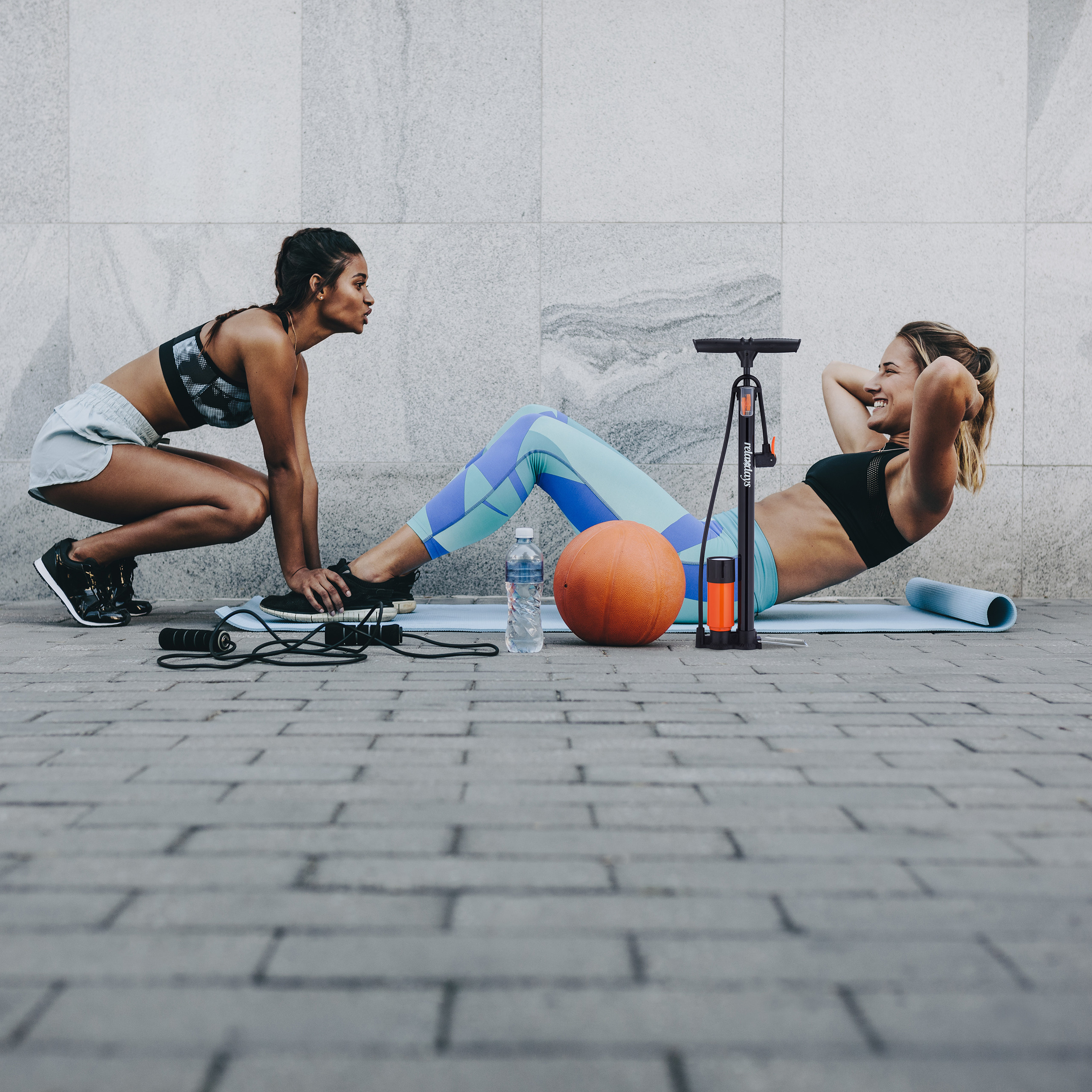 Zwei Frauen trainieren im Freien, eine macht Crunches, während die andere ihre Füße hält, mit Ball und Pumpe.