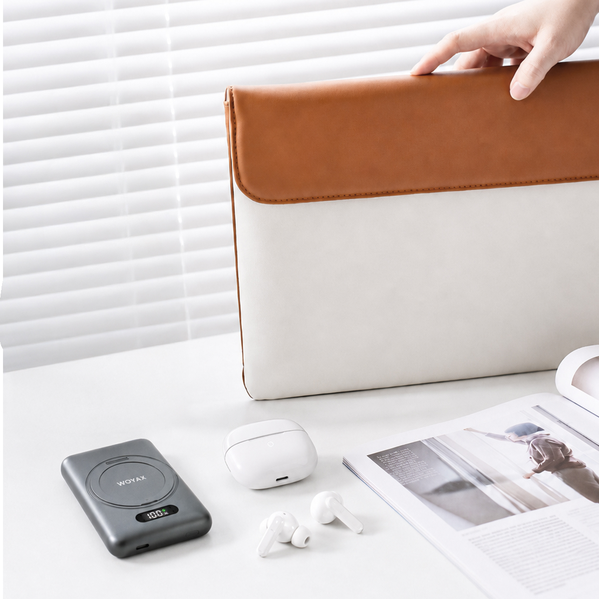 Bir el, kulaklık ve powerbank'in yanındaki masada duran beyaz ve kahverengi laptop çantasına uzanıyor.