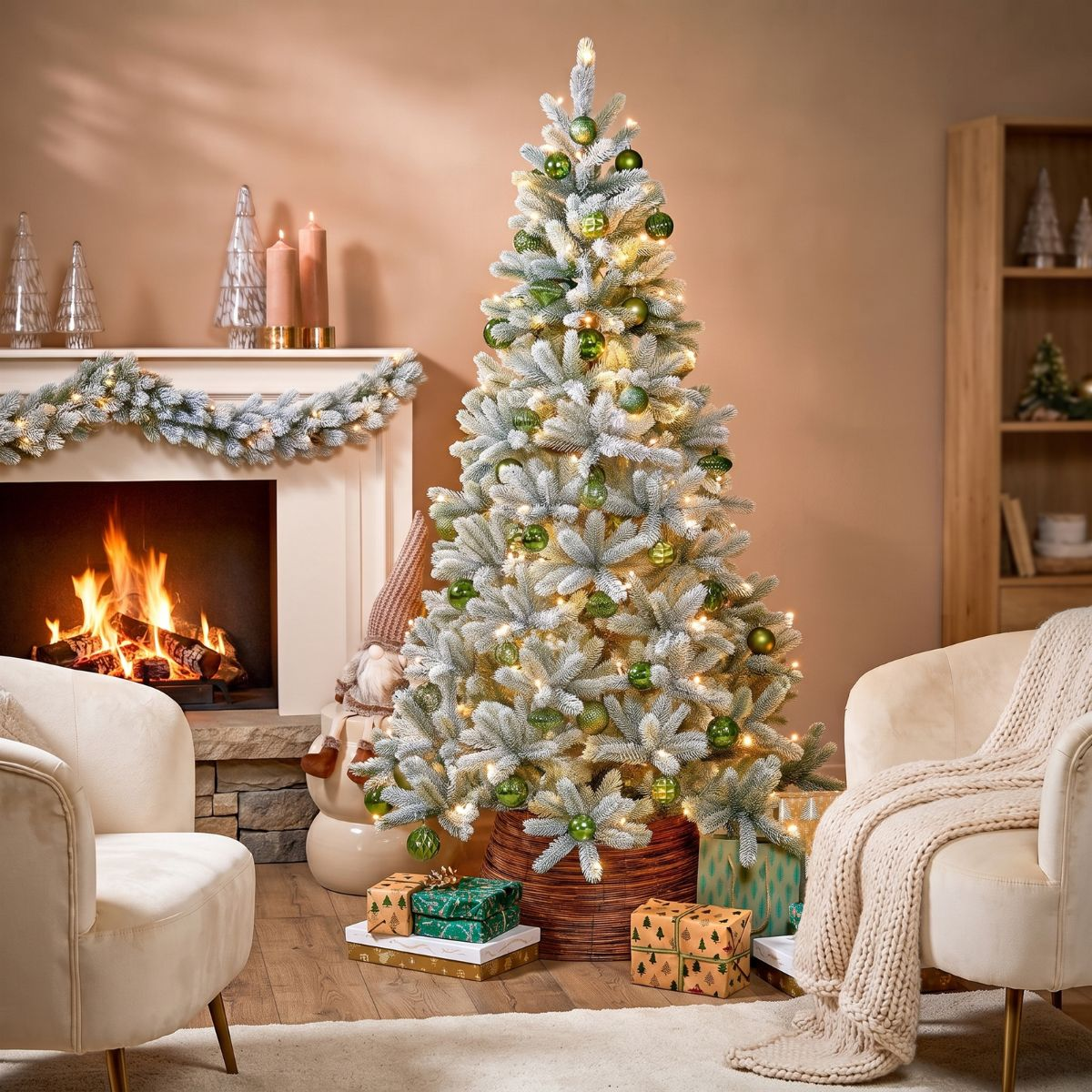 A snowy Christmas tree decorated with green ornaments and lights, positioned next to a lit fireplace and armchairs.