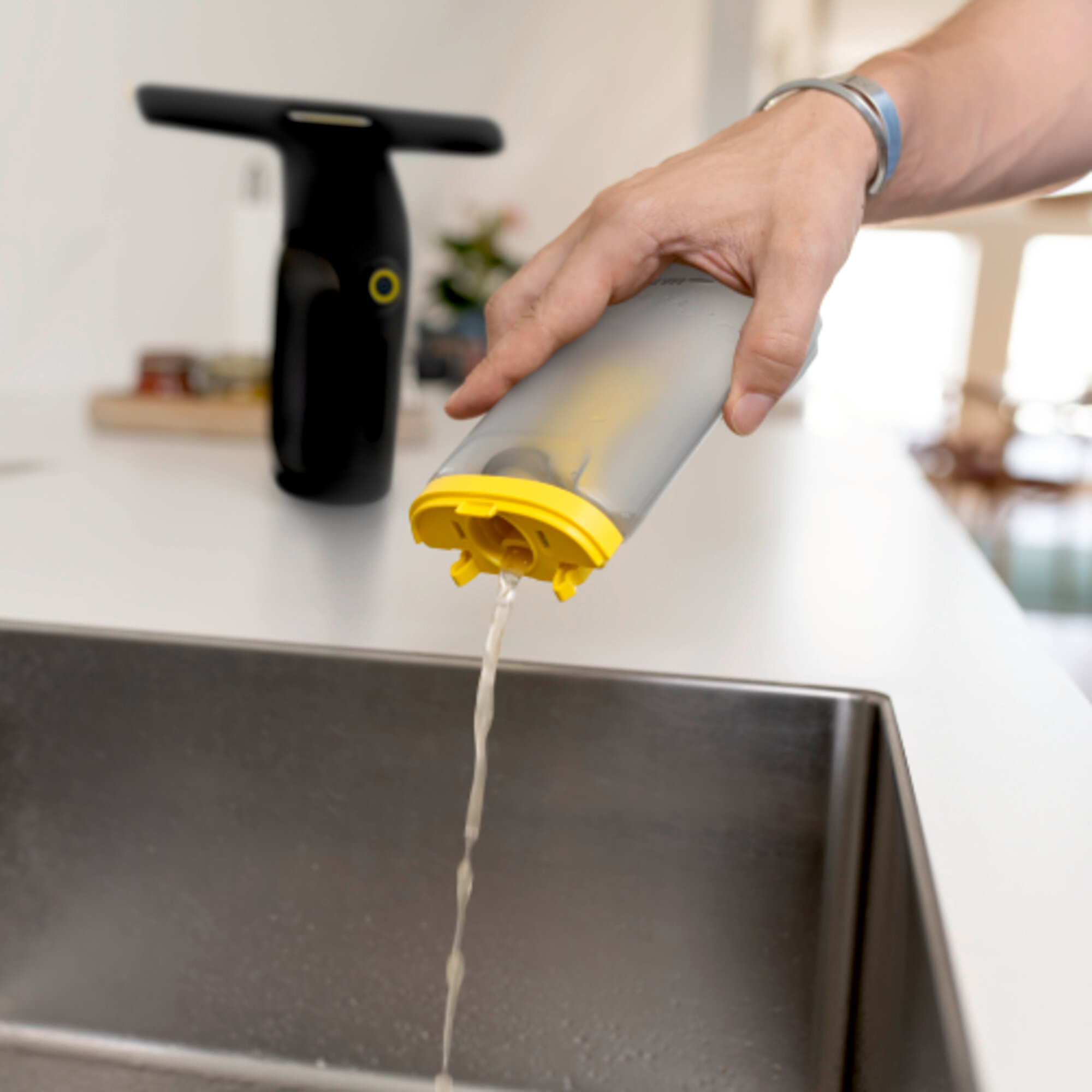 Eine Hand gießt Wasser aus einem grauen Behälter mit gelbem Deckel in ein Edelstahl-Spülbecken.