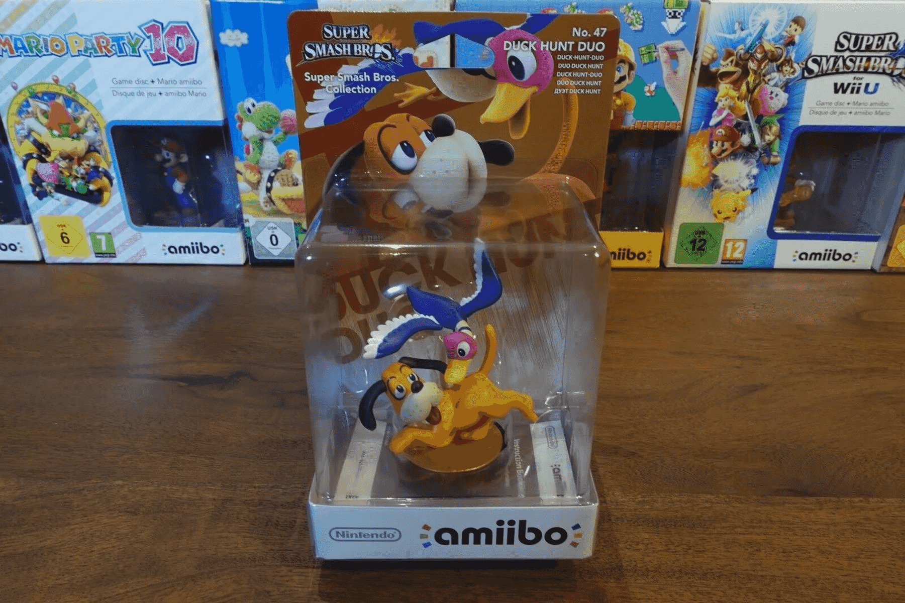 Eine Duck Hunt Duo Amiibo-Figur in ihrer Originalverpackung, eingerahmt von anderen Amiibo-Schachteln auf Holz.