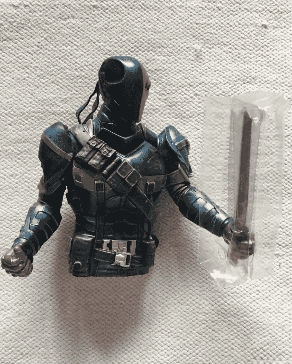 Schwarze Deathstroke-Actionfigur mit Maske, Rüstung und einem in Plastik verpackten Schwert vor weißem Hintergrund.