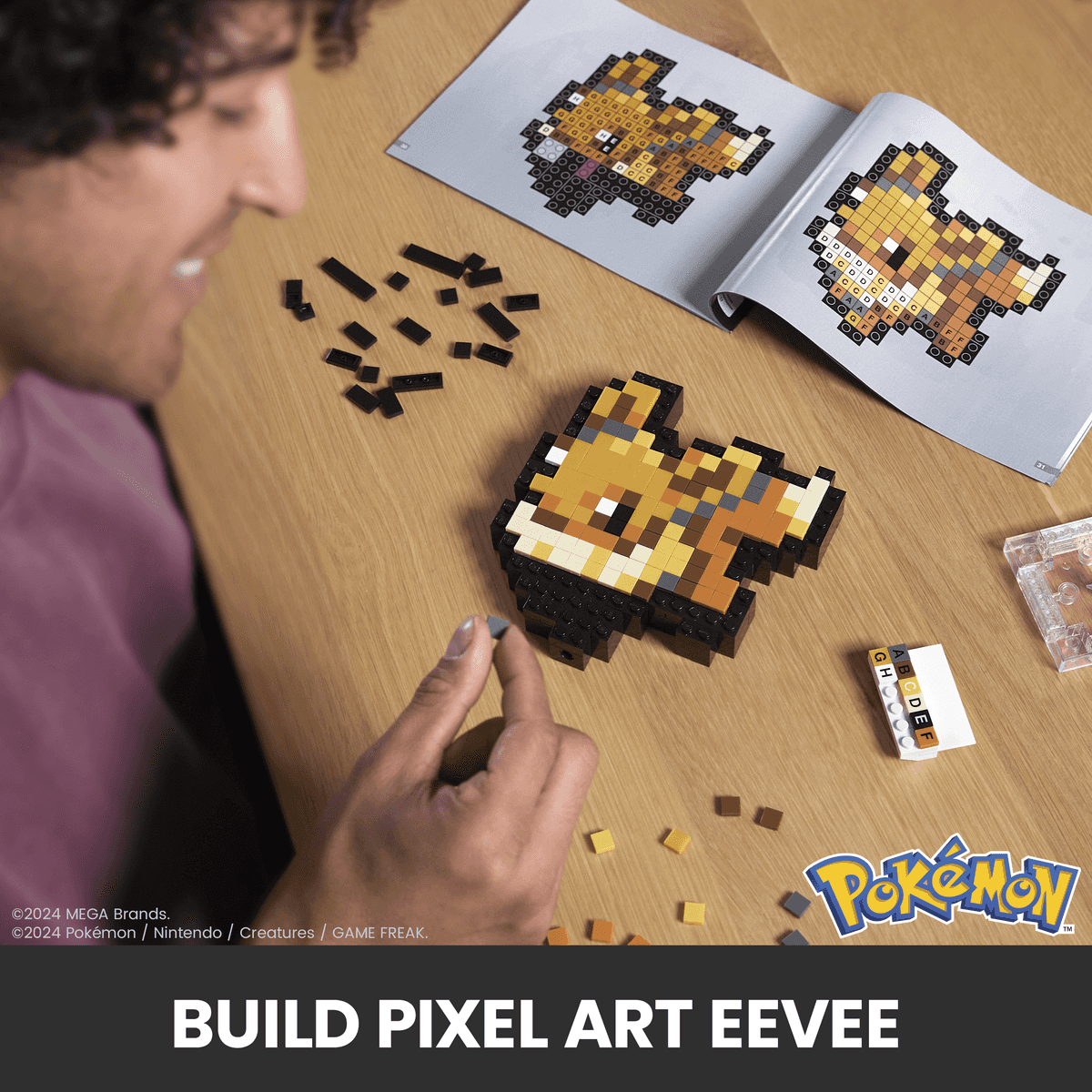 Eine lächelnde Person baut Pixel-Art-Eevee aus kleinen Steinen nach Anleitung auf einem Holztisch.