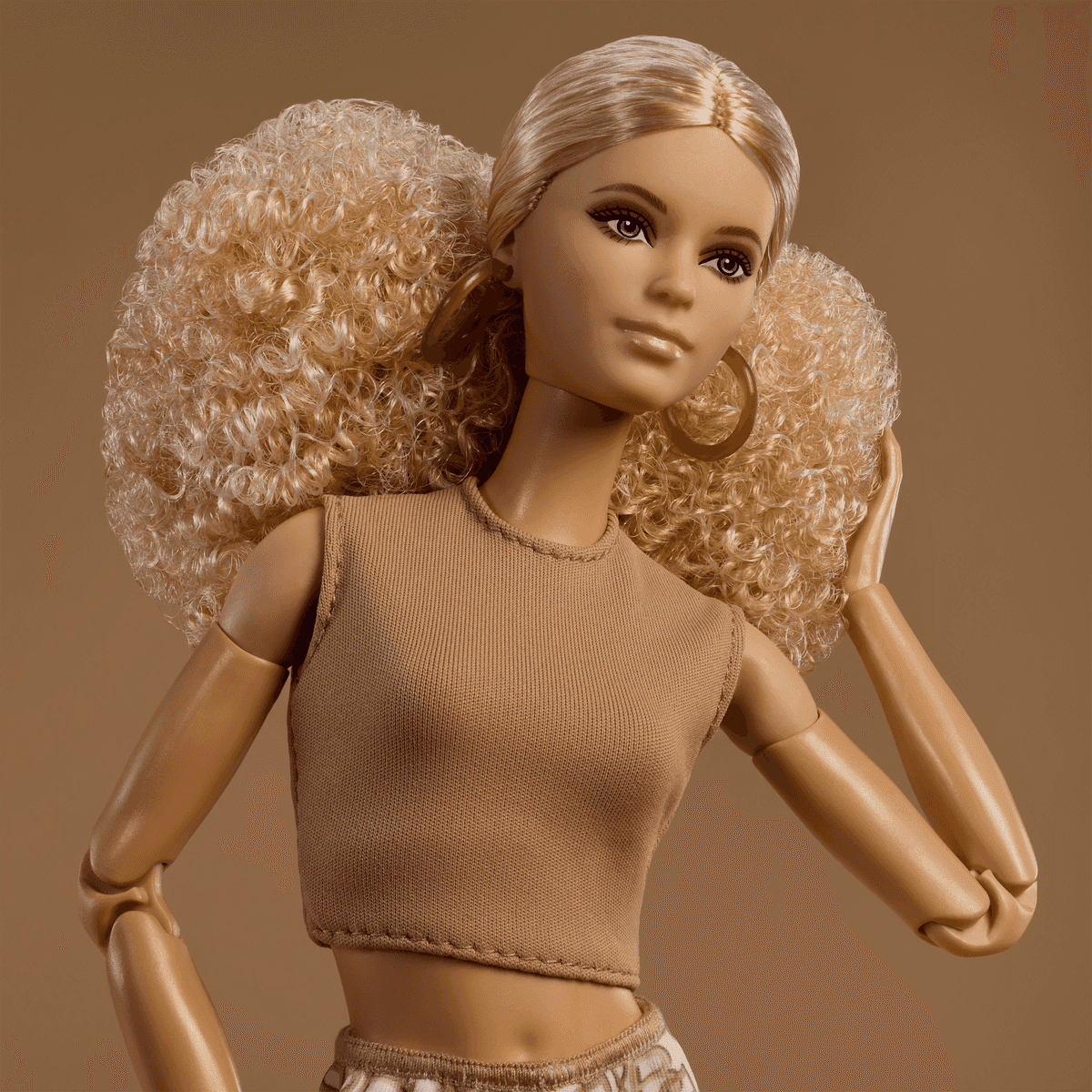 Eine blonde Fashion-Puppe mit voluminösen Locken, goldenen Creolen und einem beigen Crop-Top vor braunem Hintergrund.