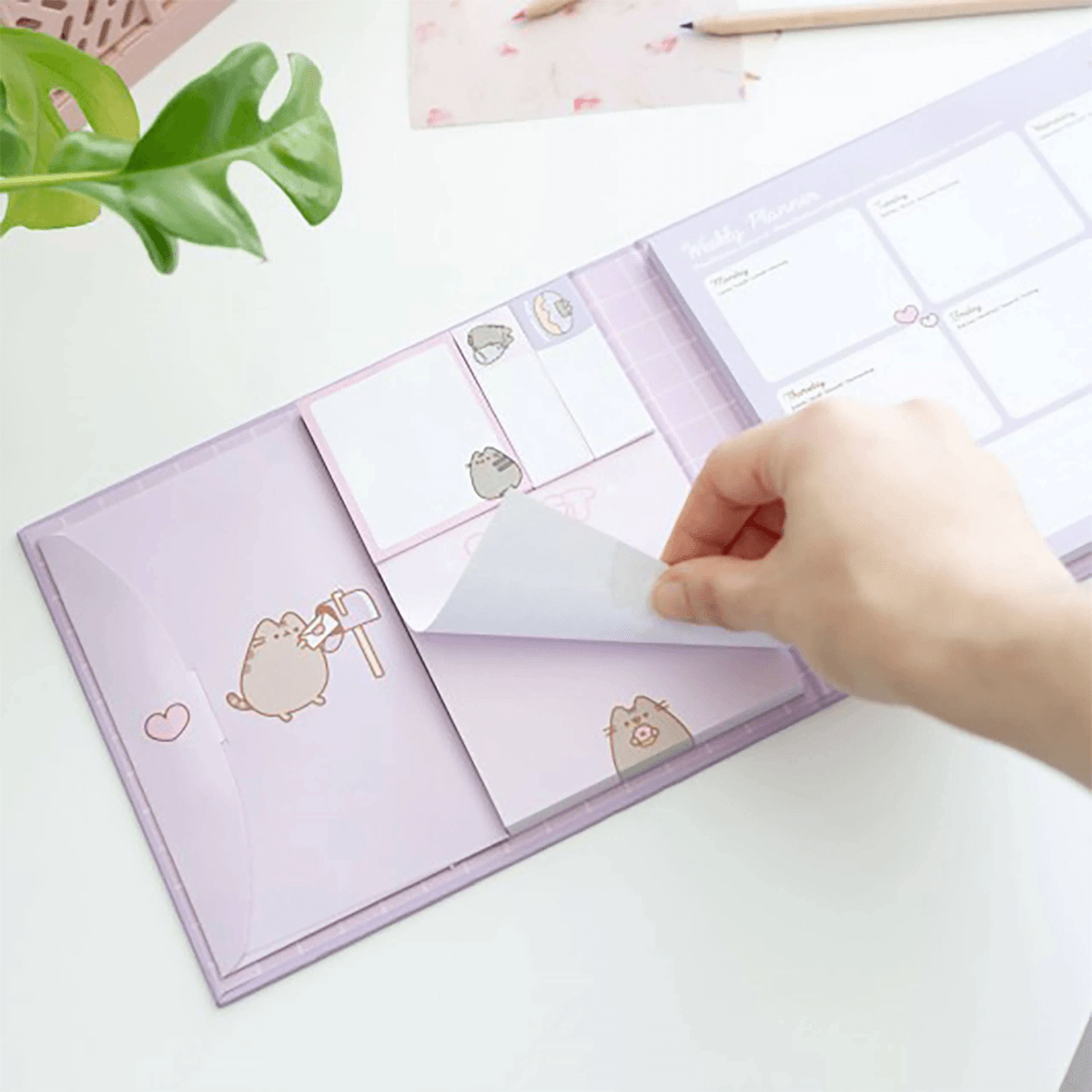 Hand blättert in einem pastelllila Notizbuch mit Katzenmotiven auf einem weißen Tisch.