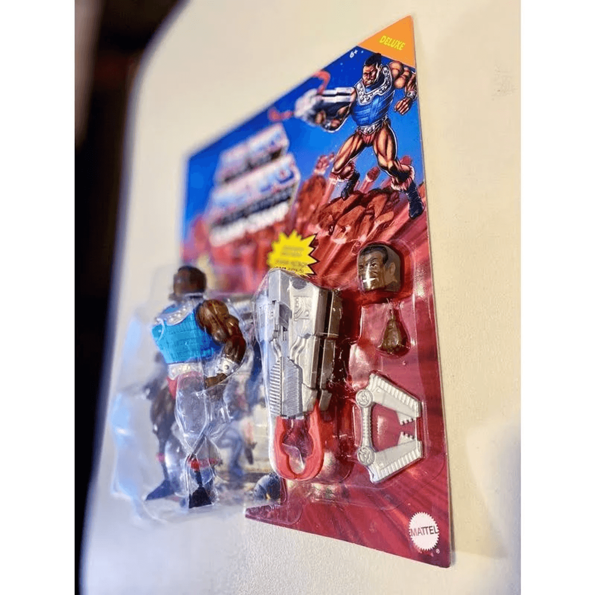 Actionfigur in Verpackung mit blauer Weste, silberner Waffe, Zusatzkopf und Zubehör auf rotem Hintergrund.