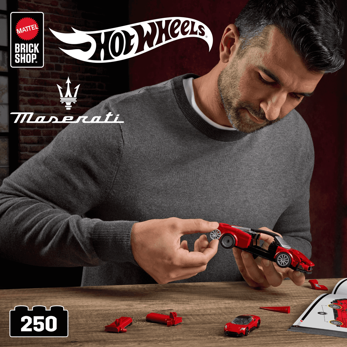 Un hombre con jersey gris monta un coche de juguete rojo en una mesa de madera, con logotipos de Hot Wheels y Maserati.
