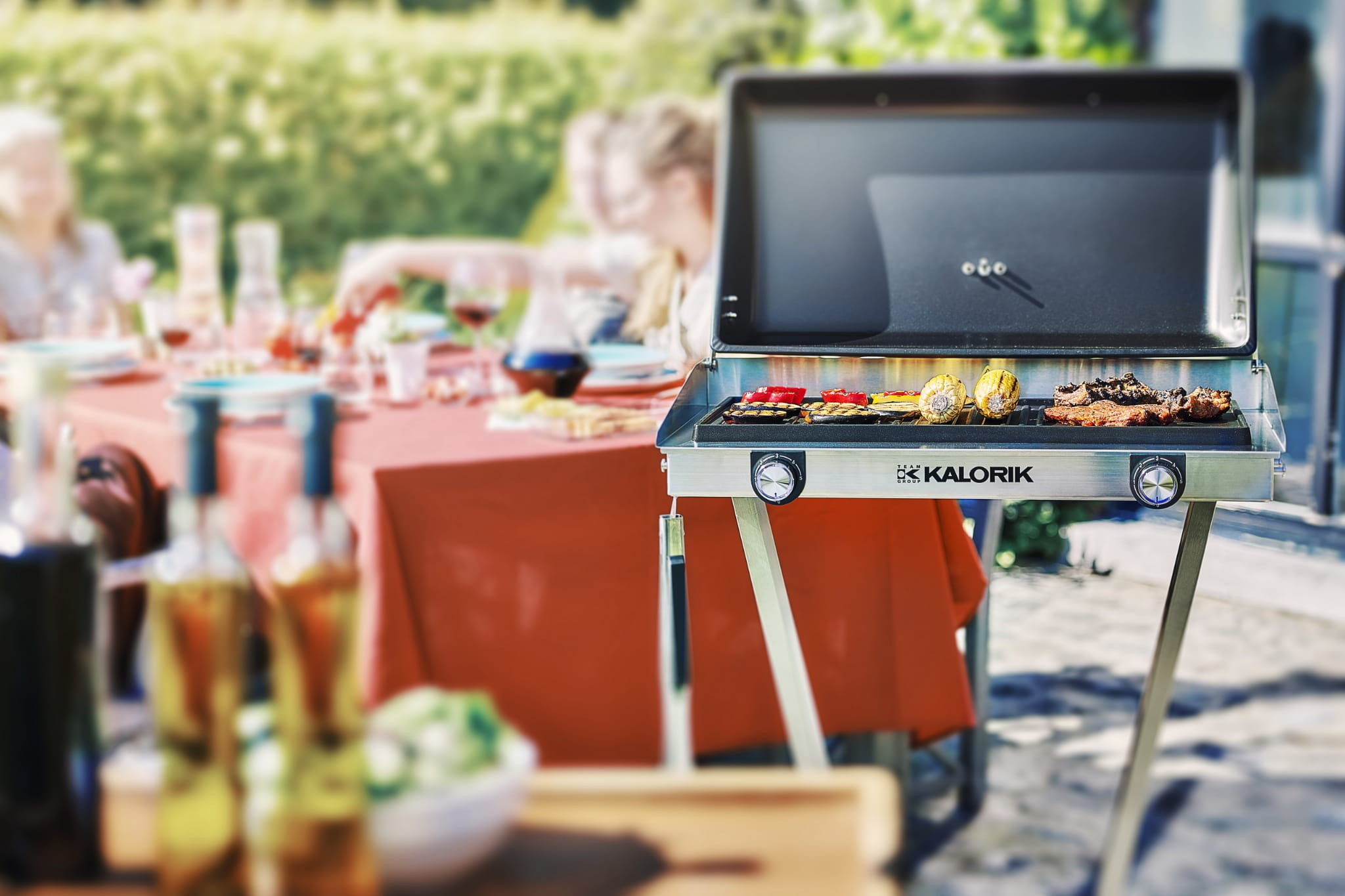 Gazowy grill Kalorik z jedzeniem stoi na zewnątrz, a w tle widać rozmytą grupę ludzi.