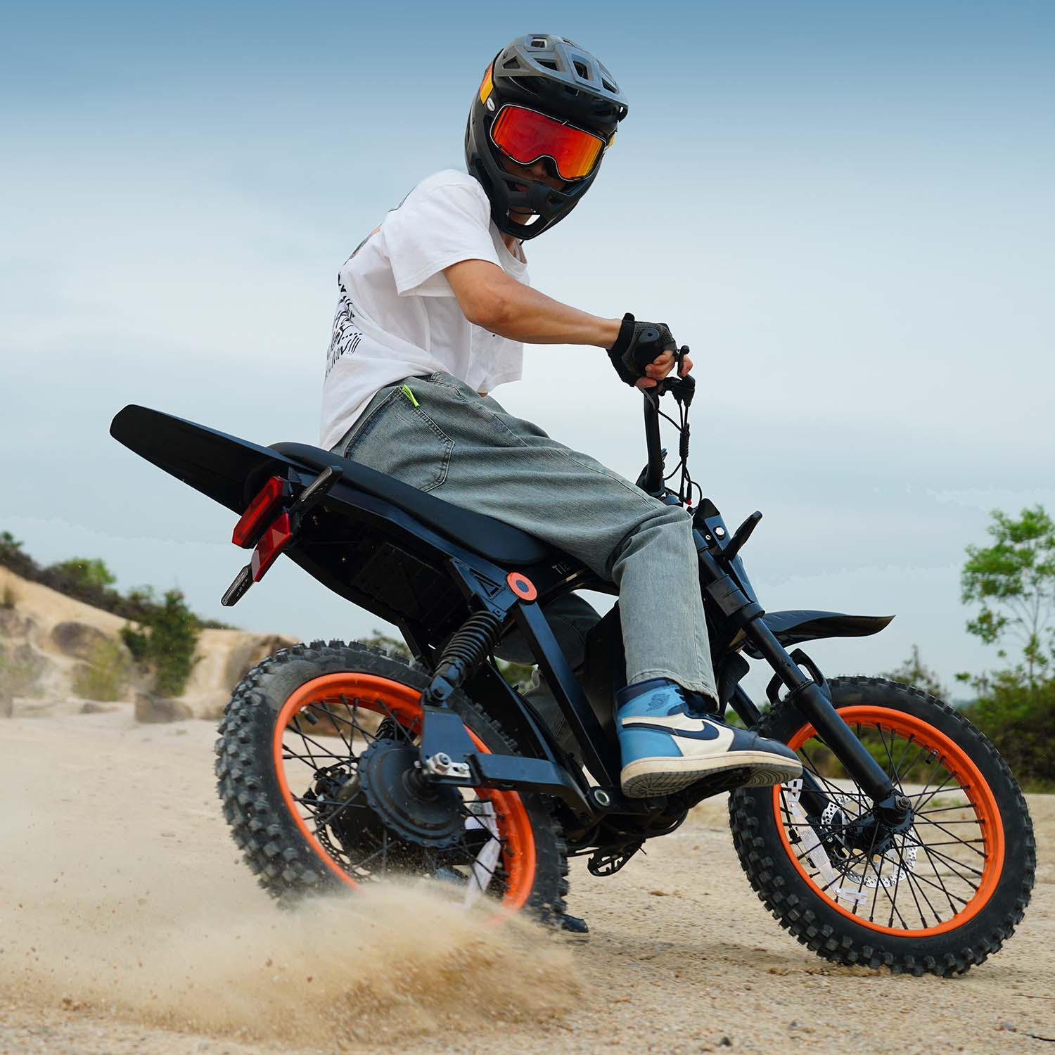 Kierowca w kasku i pomarańczowych goglach jedzie czarnym motocyklem elektrycznym po piasku.