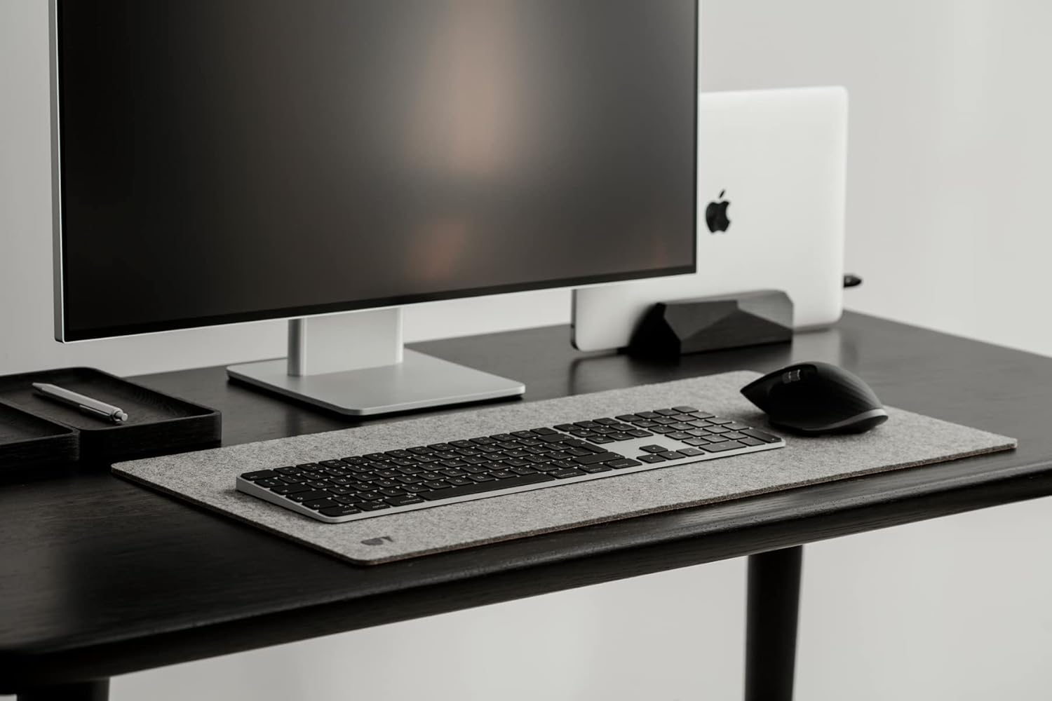 Nowoczesne biurko z monitorem, zamkniętym laptopem, klawiaturą i myszką na szarej filcowej podkładce.