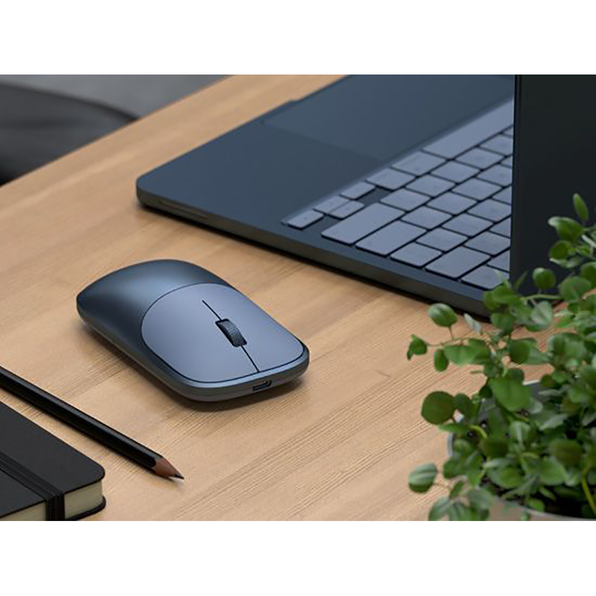 Eine blaue Computermaus auf einem Holztisch neben einem Laptop, Stift, Notizbuch und einer kleinen Topfpflanze.