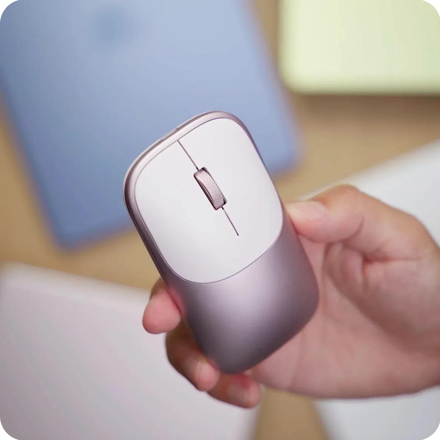 Eine Hand hält eine schlichte kabellose Computermaus mit rosa metallischem Körper und weißer Oberseite über einem Schreibtisch.