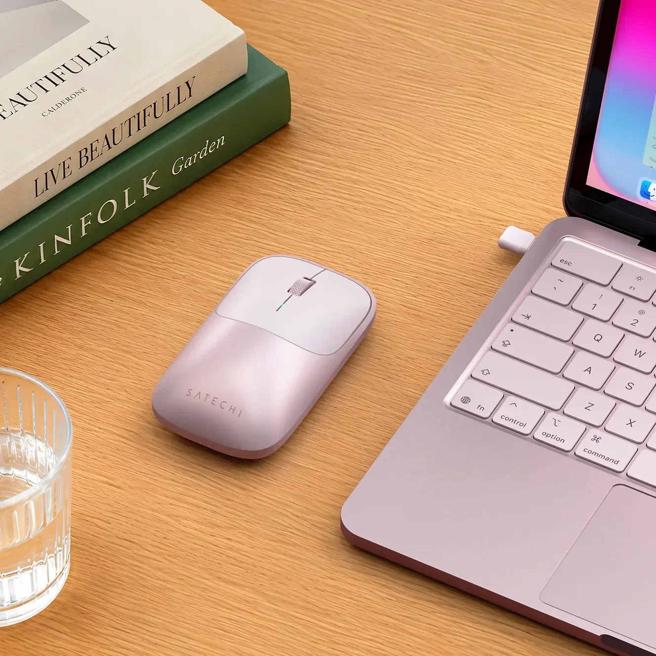 Eine rosa Satechi-Maus auf einem Holztisch, neben einem Laptop, Büchern und einem Glas Wasser.