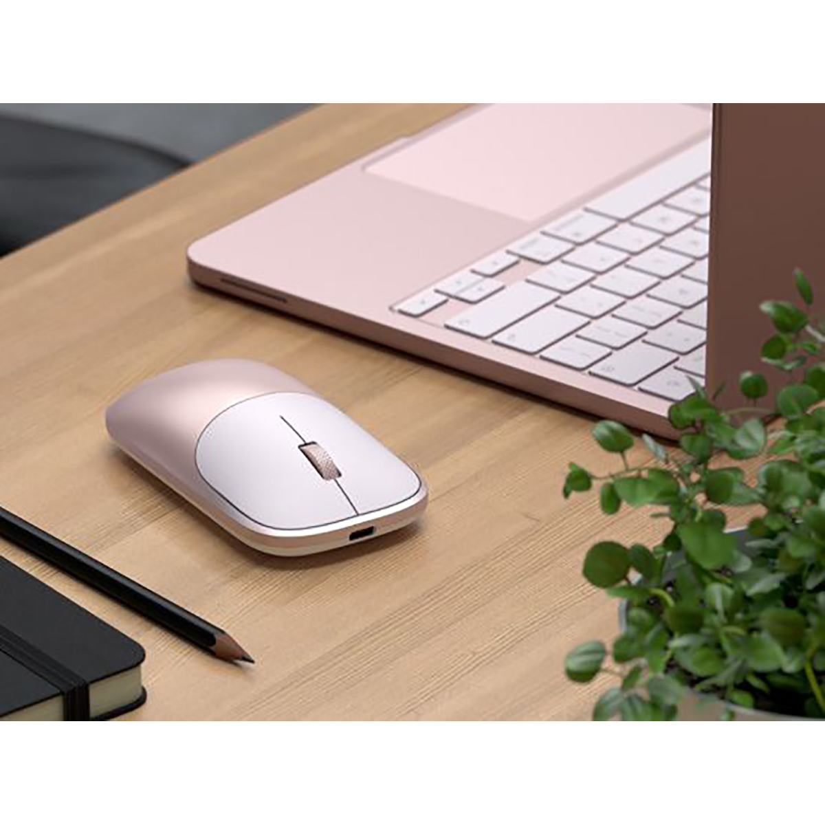 Eine rosafarbene und weiße Computermaus auf einem Holztisch, neben einem Laptop, einem Stift und einer Pflanze.