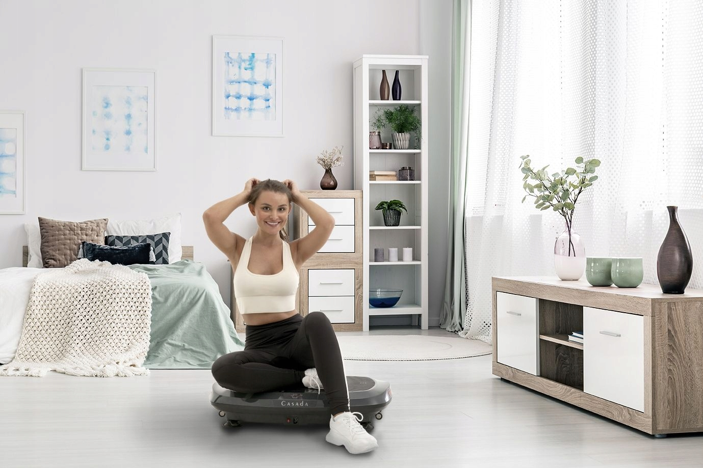 Uśmiechnięta kobieta w stroju sportowym siedzi na platformie wibracyjnej w jasnym pokoju.