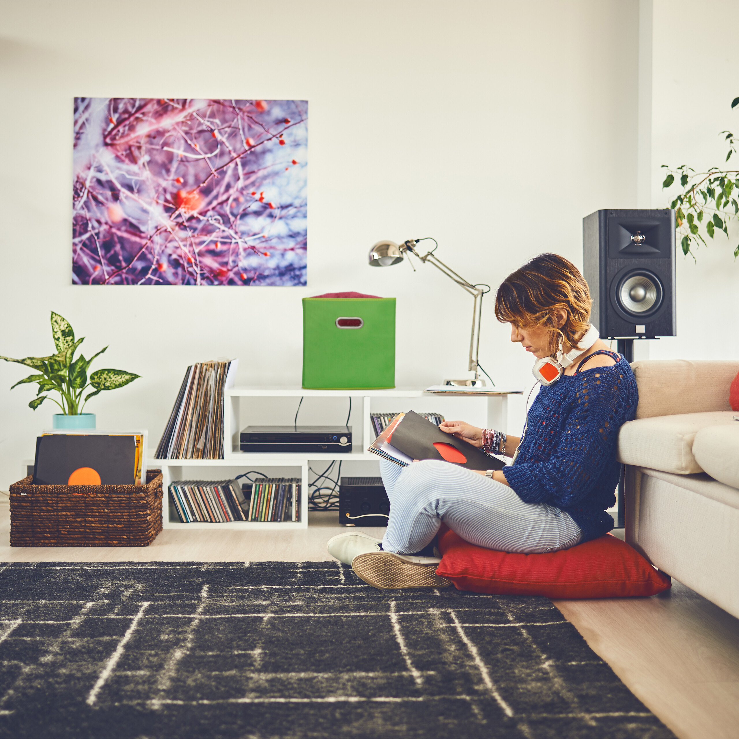 Eine Frau sitzt auf einem roten Kissen am Boden und sortiert Vinyl-Schallplatten in einem hellen Wohnzimmer.