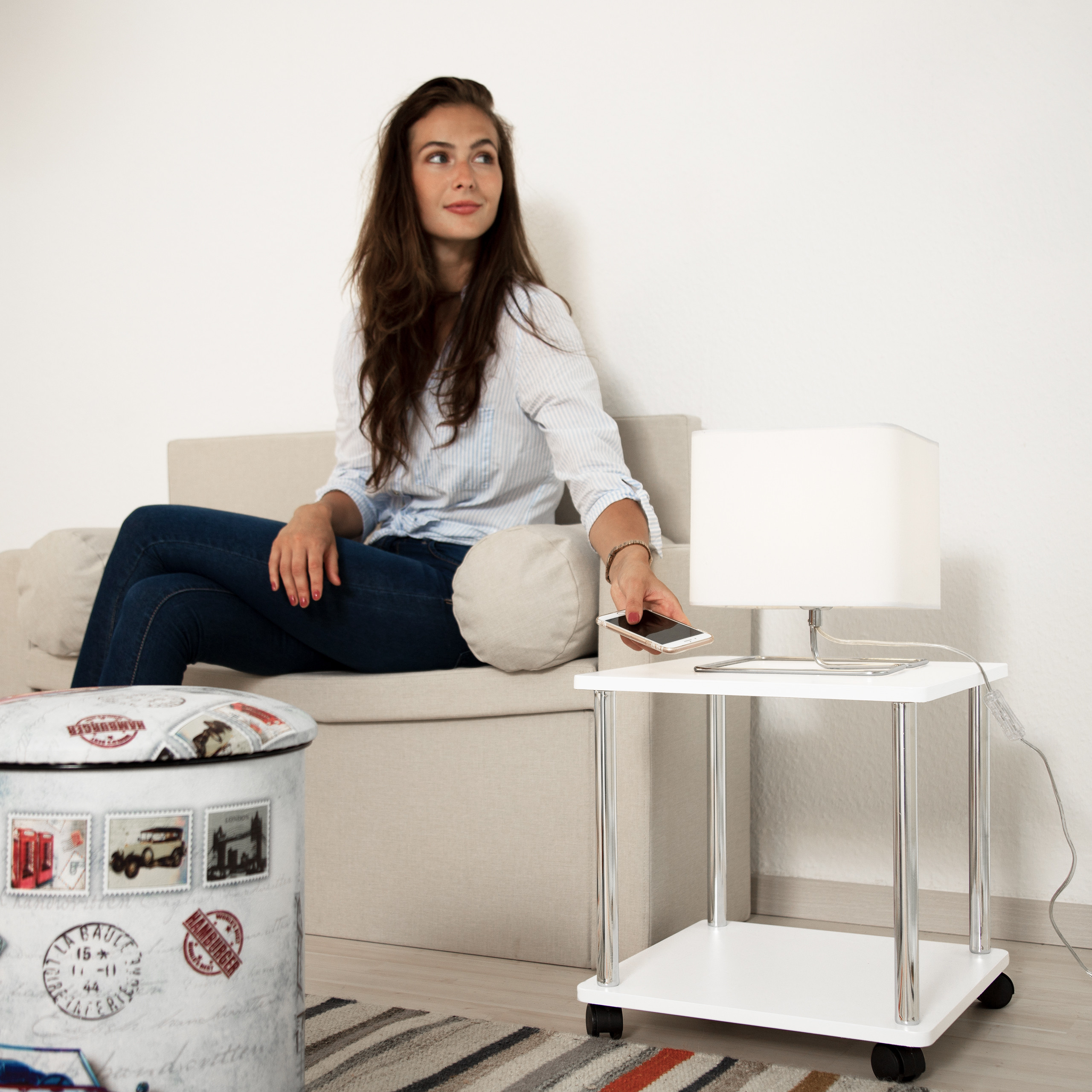 Eine Frau sitzt auf einem hellen Sofa und legt ihr Smartphone auf einen kleinen Tisch mit Lampe.
