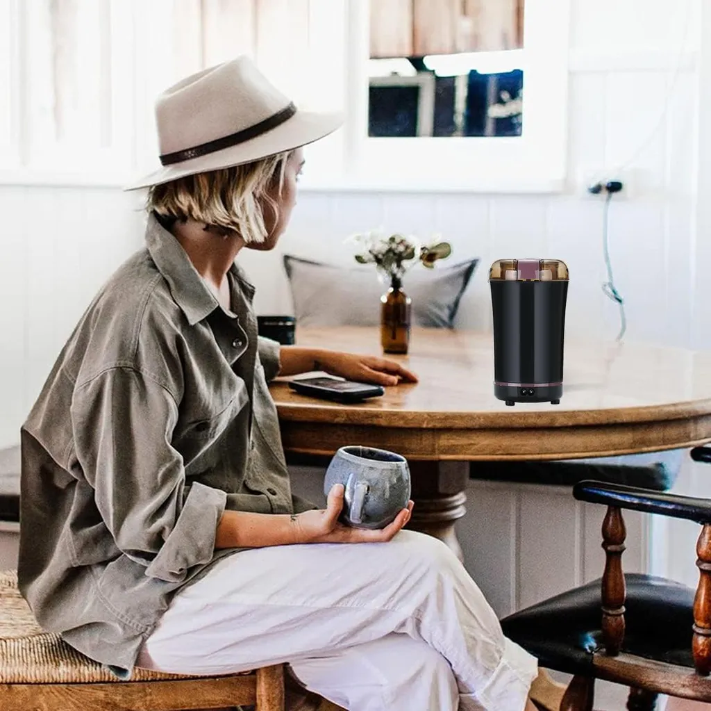 Frau mit Hut und Hemd sitzt am Tisch und hält eine Tasse, daneben eine Kaffeemühle.