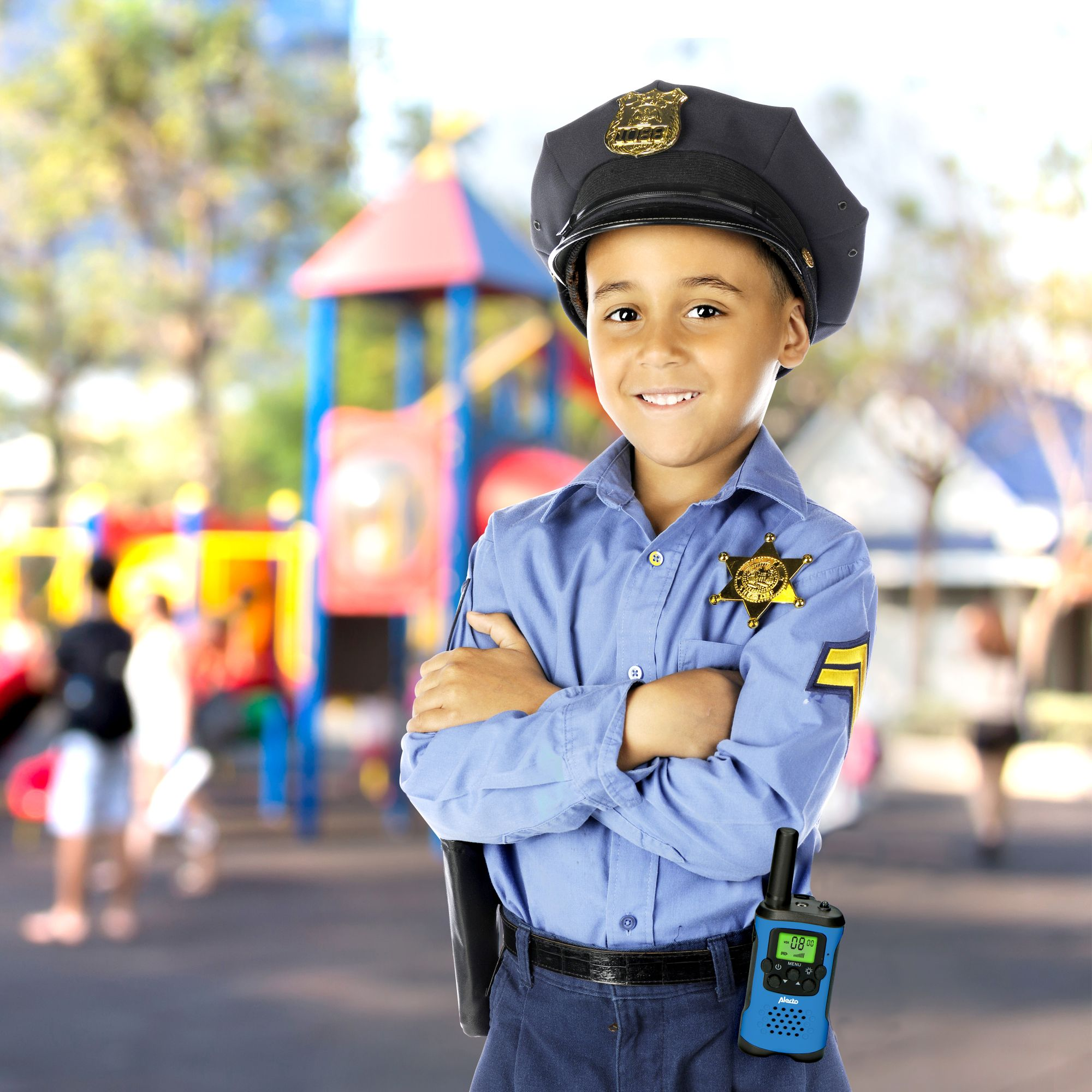 Un bambino sorridente vestito da poliziotto con walkie-talkie davanti a un parco giochi.