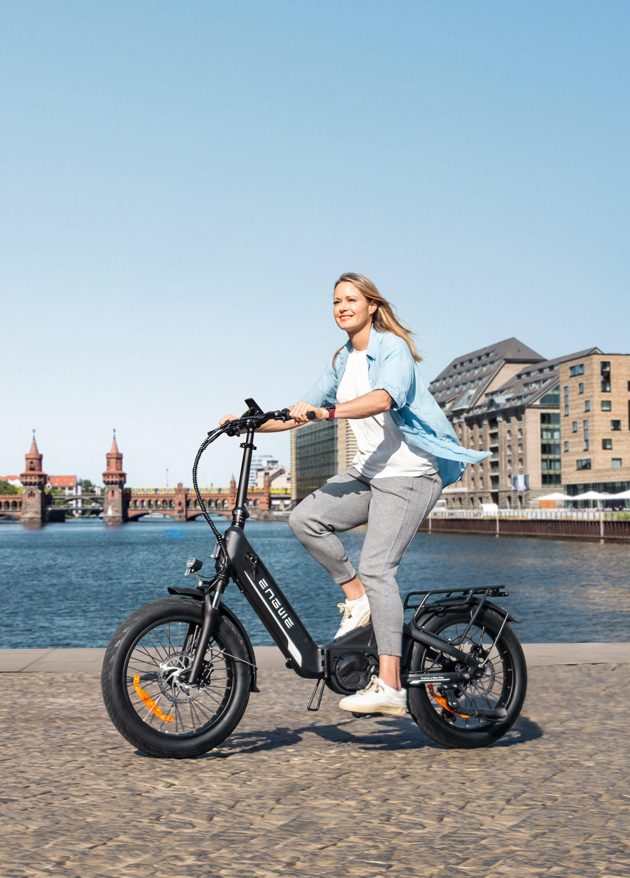 Una donna guida una bici elettrica nera lungo una passeggiata sul fiume con edifici storici sullo sfondo.