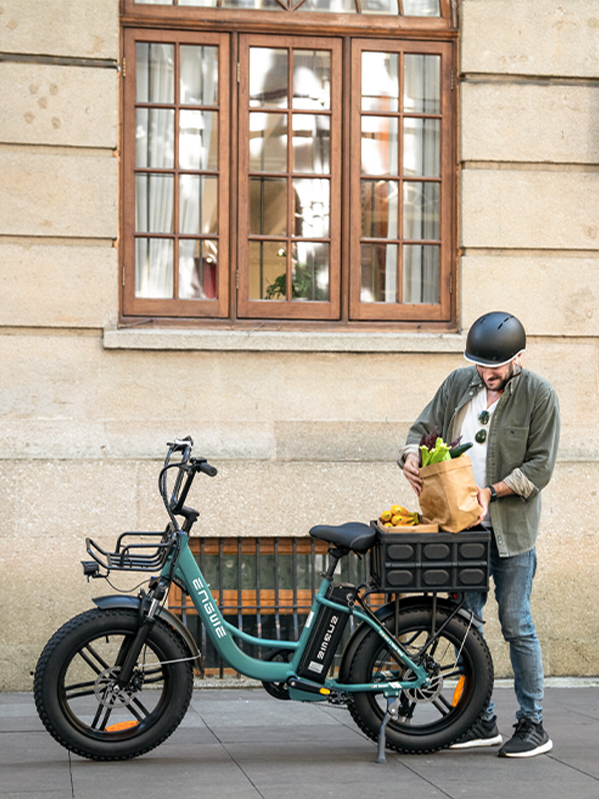 Un uomo con casco carica borse della spesa su una e-bike verde davanti a un muro.
