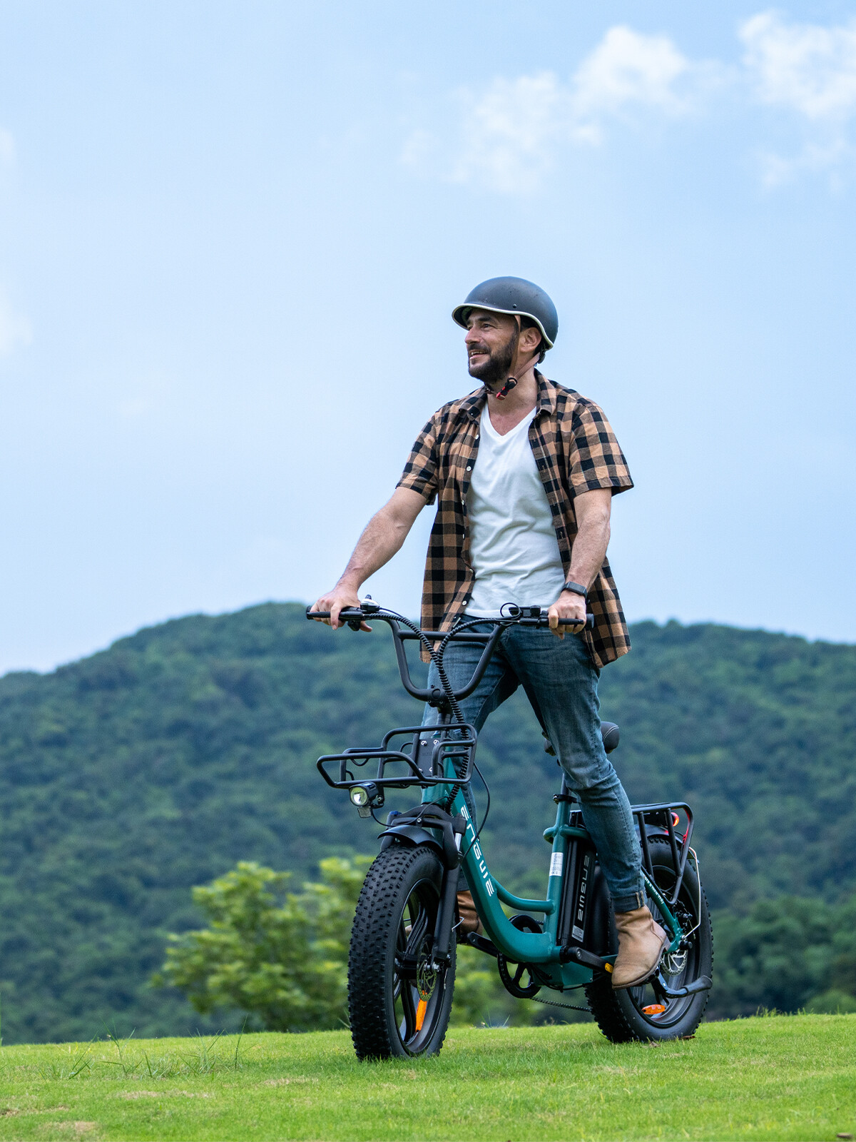 Un uomo sorridente con casco e camicia a quadri guida una e-bike verde acqua su una collina erbosa.