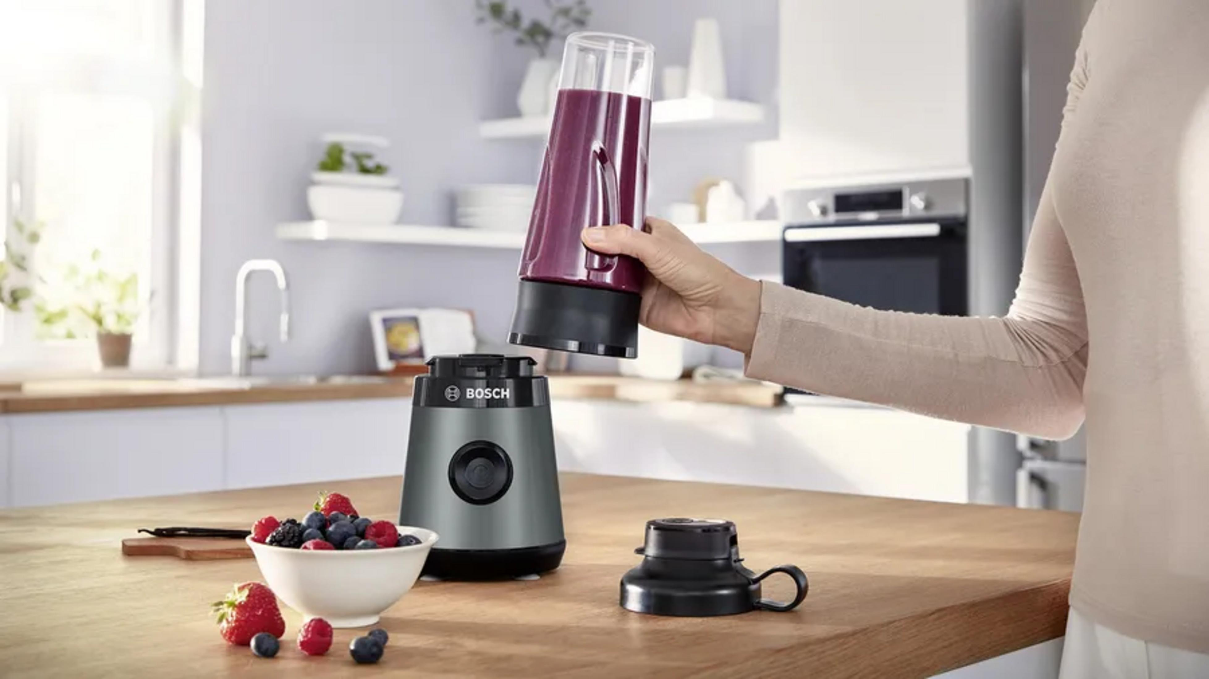 Une femme tient un gobelet de blender rempli au-dessus d'une base de blender Bosch grise dans une cuisine lumineuse.