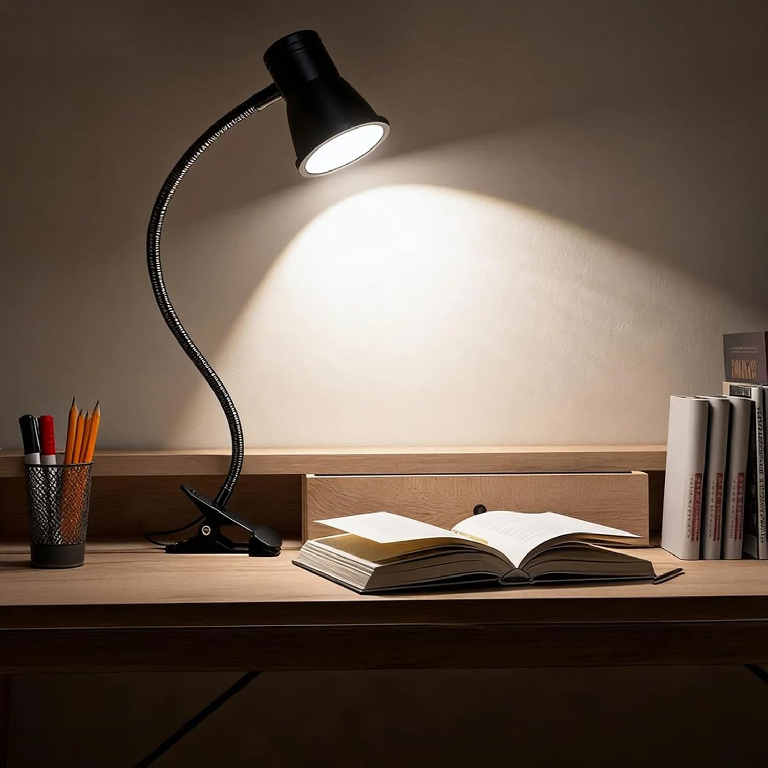 Eine brennende schwarze Schreibtischlampe beleuchtet ein offenes Buch auf einem Holztisch mit Stiften und Büchern.