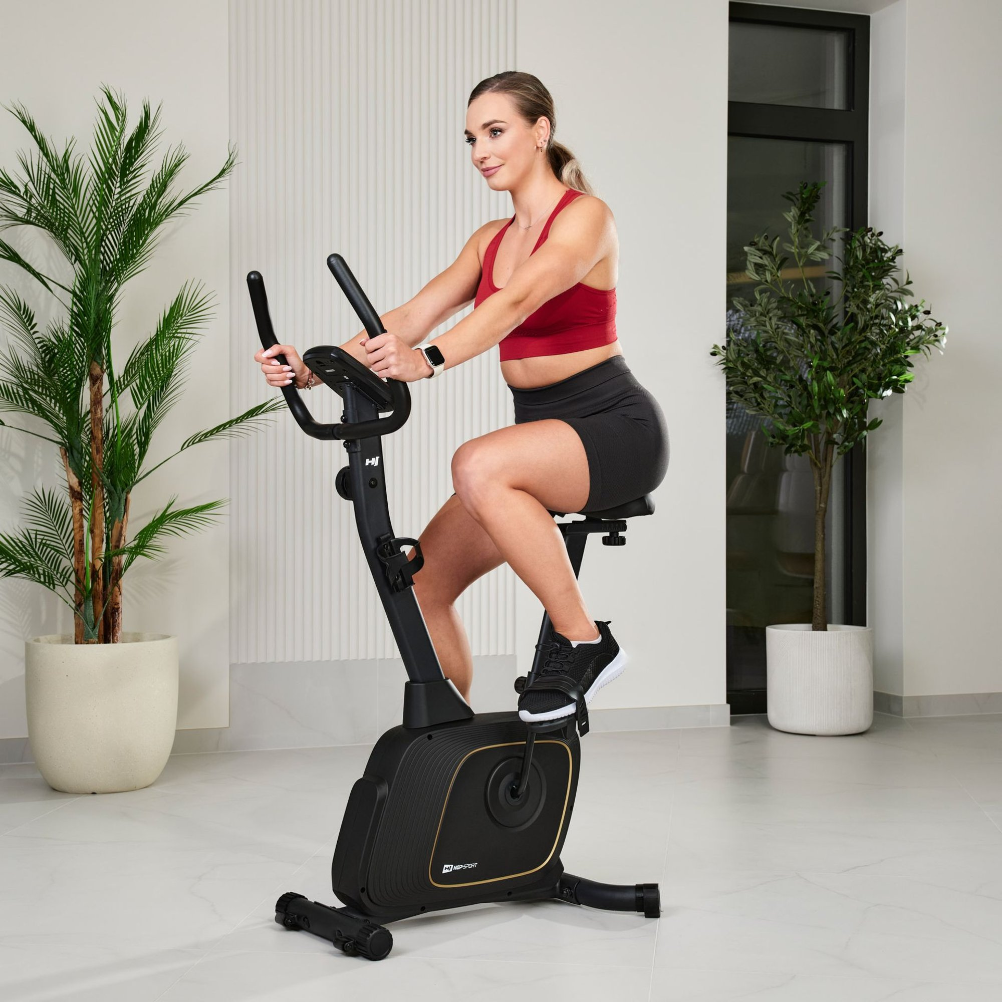 Eine Frau in rotem Top und schwarzen Shorts trainiert drinnen auf einem schwarzen Heimtrainer mit Pflanzen im Hintergrund.