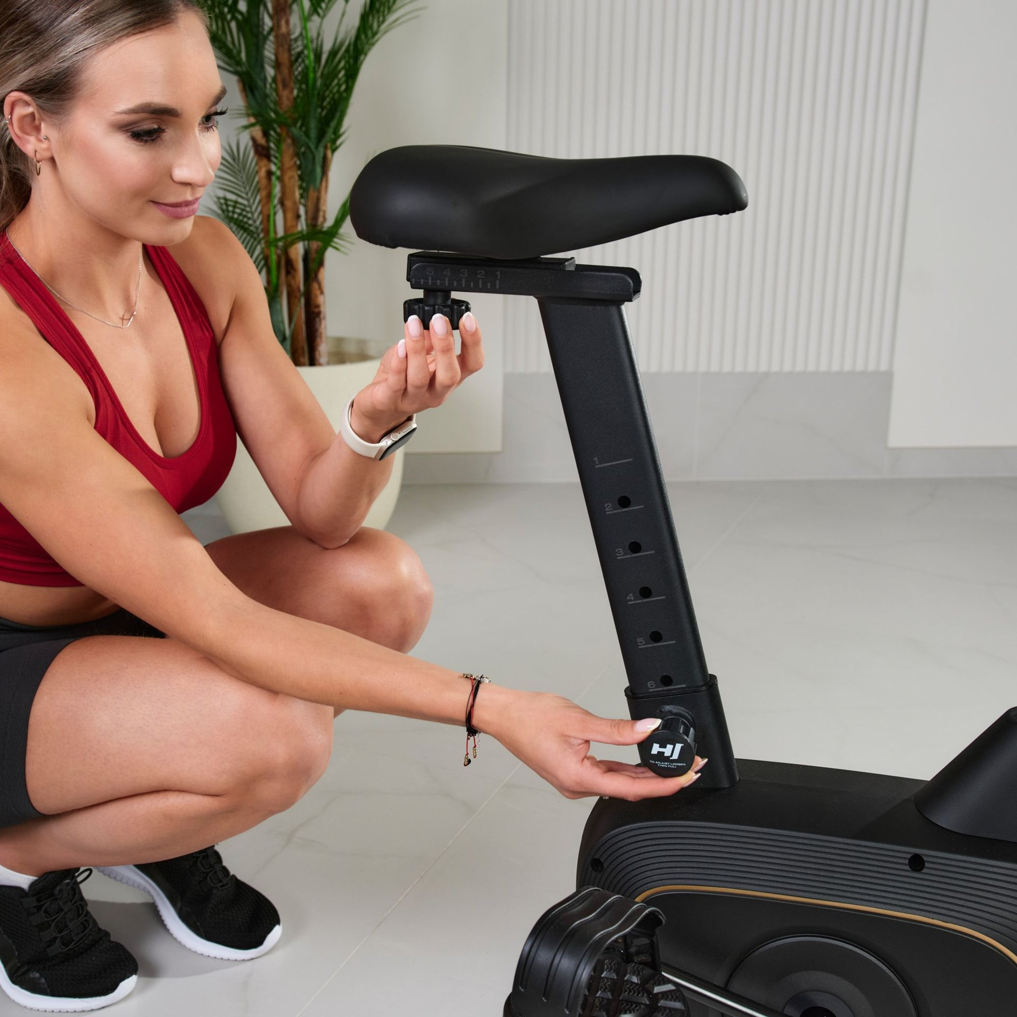 Eine Frau in Sportkleidung kniet neben einem Heimtrainer und stellt die Sitzhöhe ein.