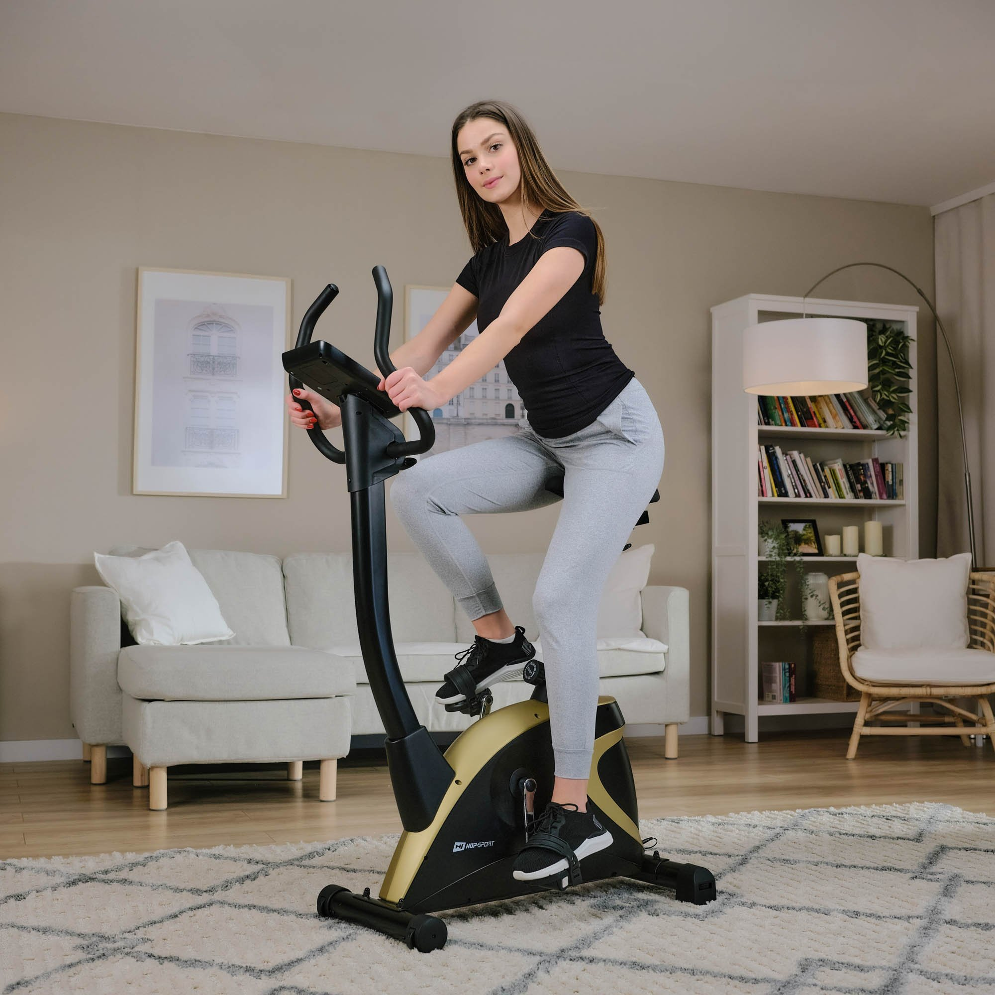 Eine Frau in grauer Jogginghose und schwarzem Shirt fährt in einem hellen Wohnzimmer Heimtrainer.