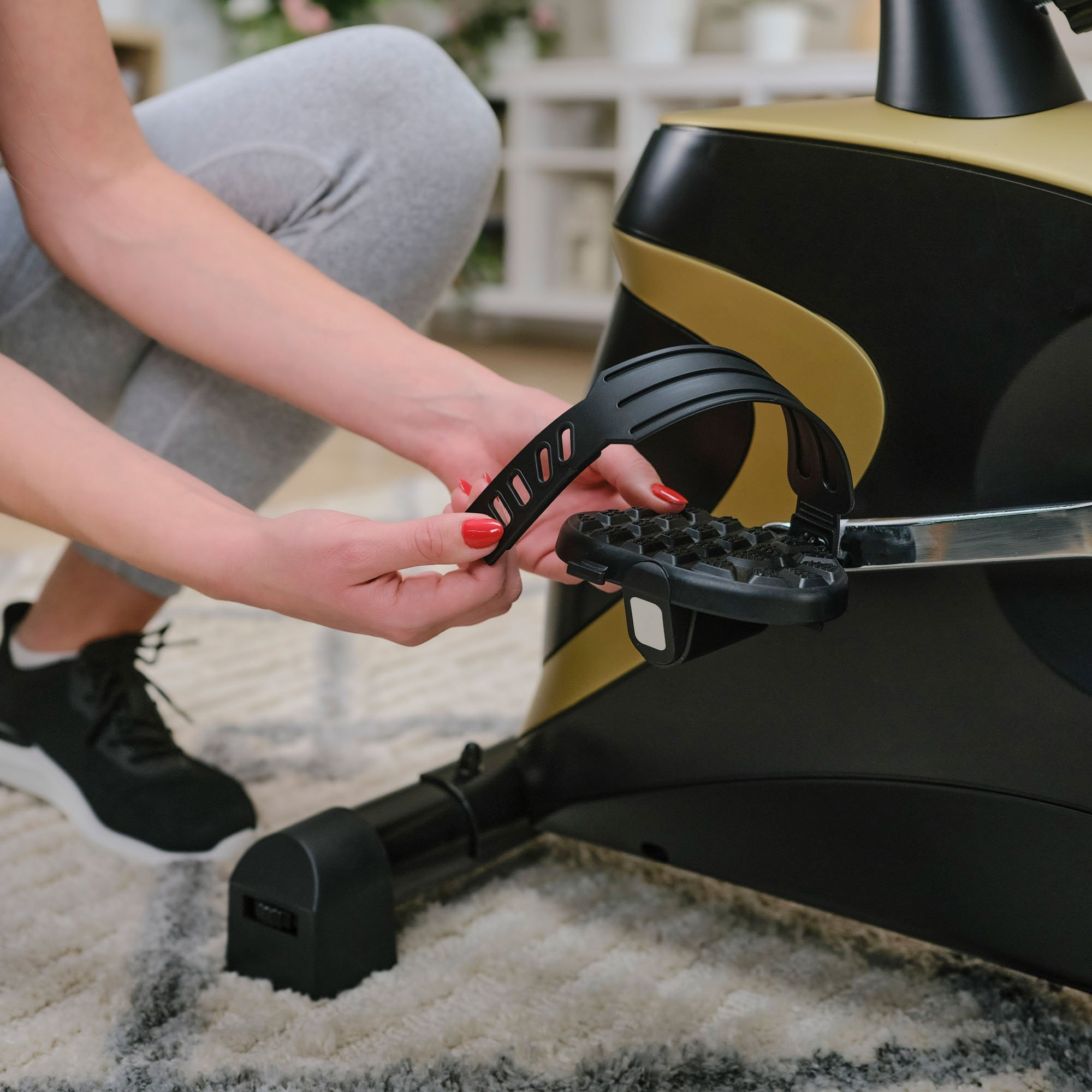 Eine Frau in grauen Leggings stellt den schwarzen Fußriemen an einem Heimtrainer ein.
