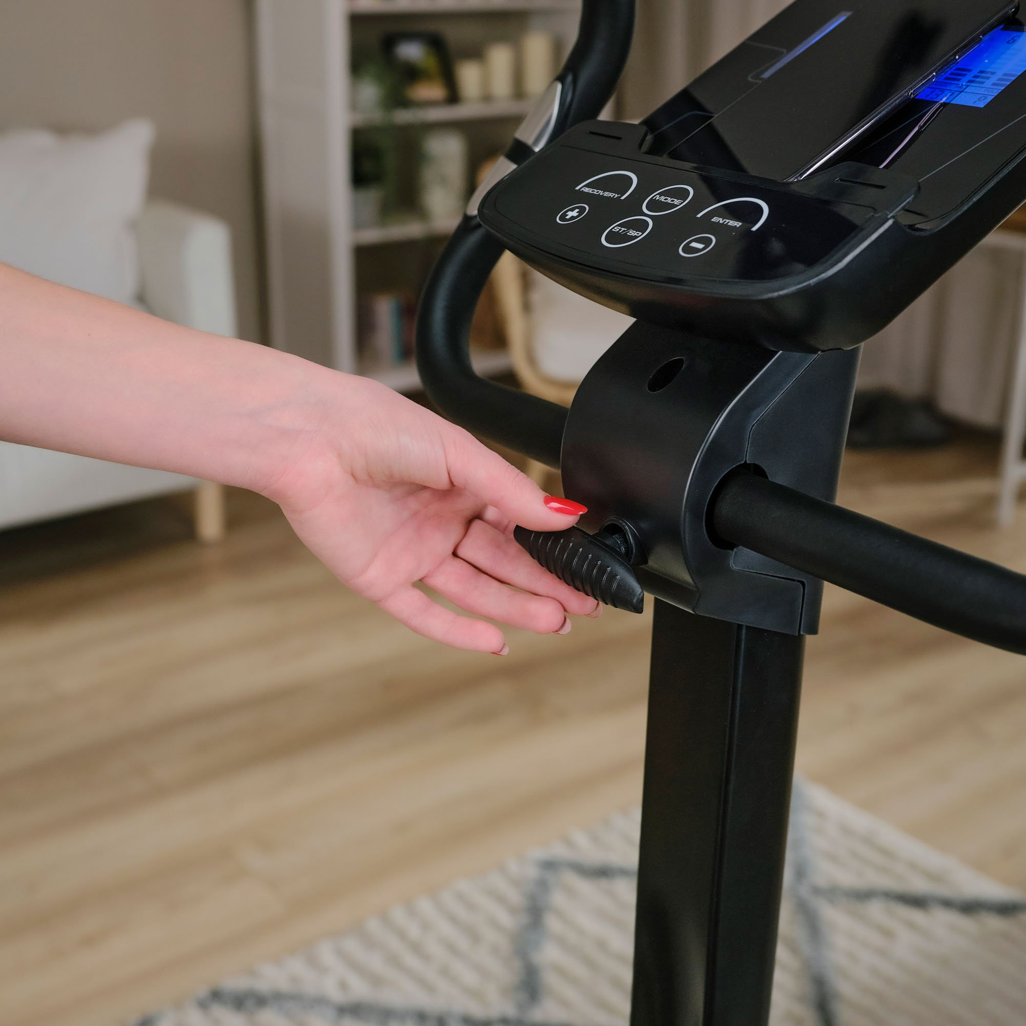 Eine Hand greift nach dem Einstellknopf an einem schwarzen Heimtrainer in einem hellen Wohnzimmer.