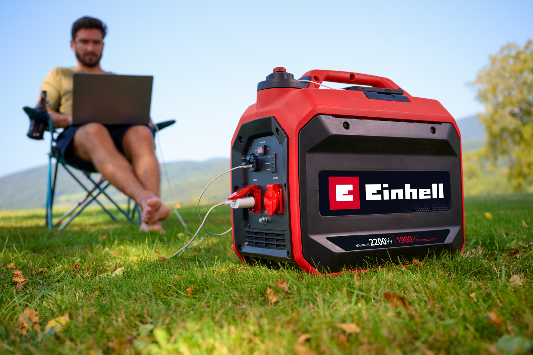 Ein roter Einhell Inverter-Stromerzeuger steht auf einer Wiese, während ein Mann im Hintergrund einen Laptop nutzt.