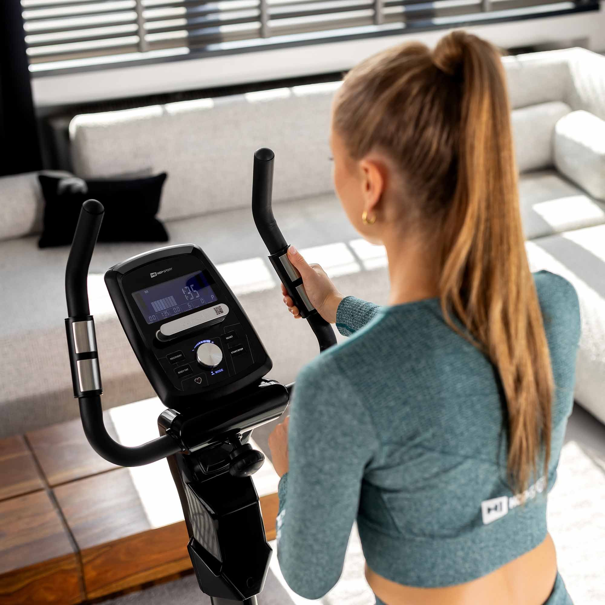 Eine Frau mit einem Pferdeschwanz trainiert auf einem Crosstrainer in einem hellen Wohnzimmer.
