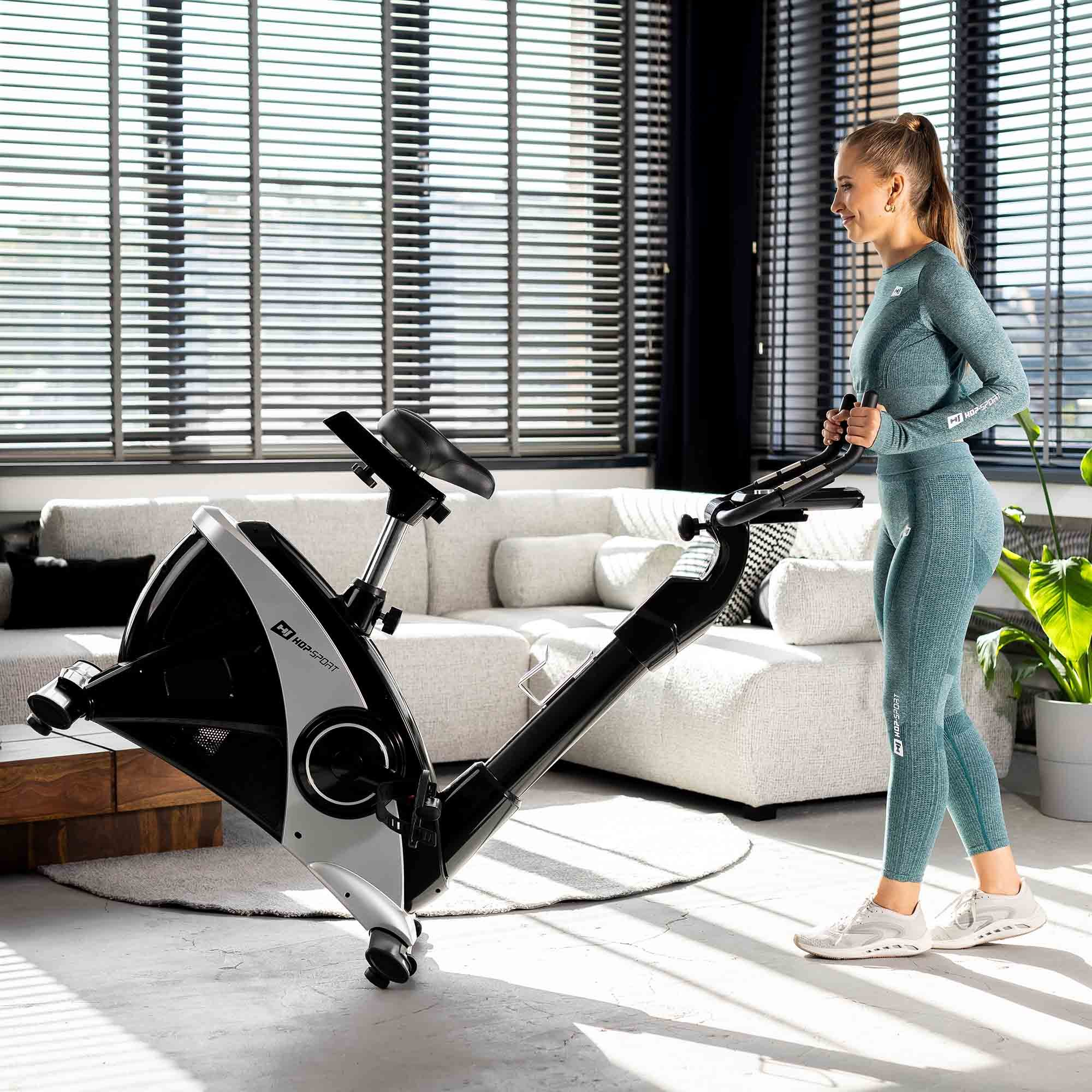 Eine Frau in Sportkleidung steht neben einem modernen Heimtrainer in einem hellen Wohnzimmer.