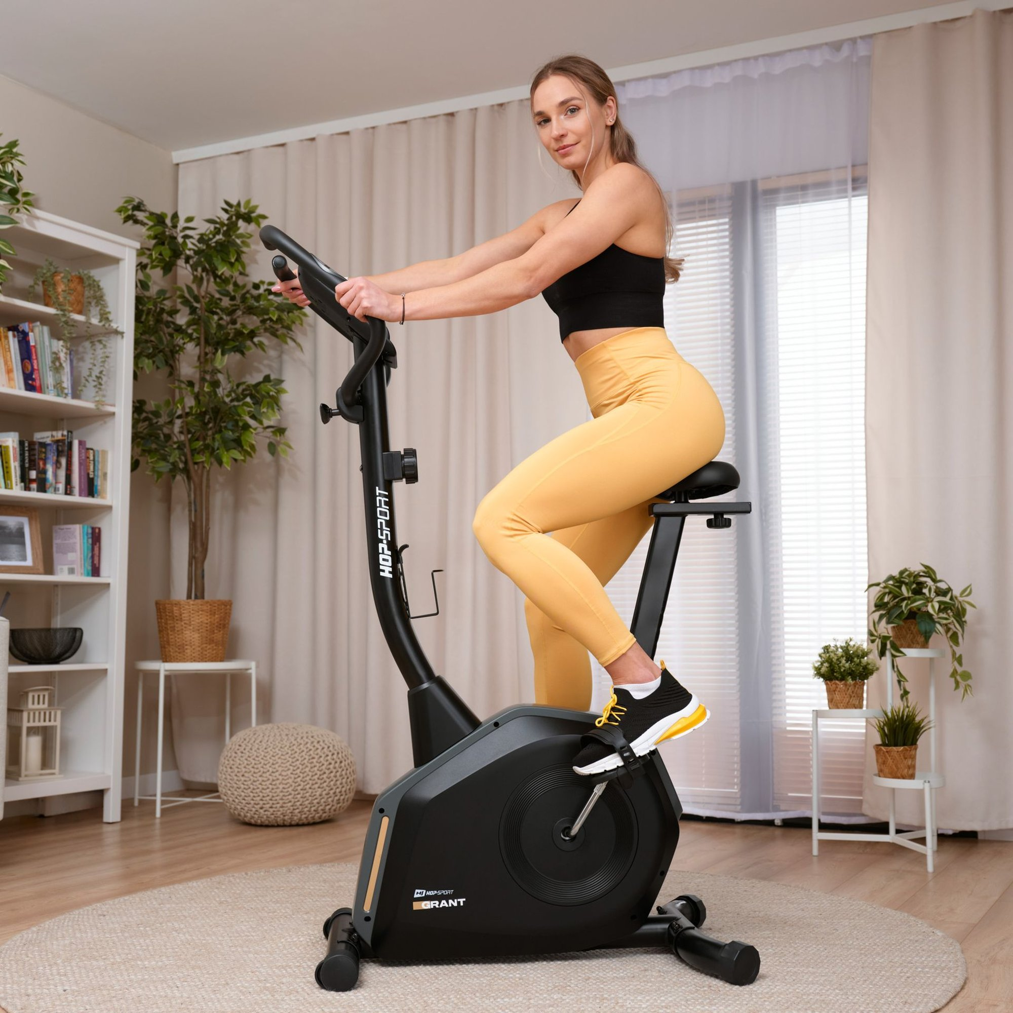 Eine Frau in gelben Leggings und schwarzem Top trainiert auf einem Heimtrainer in einem hellen Wohnzimmer.