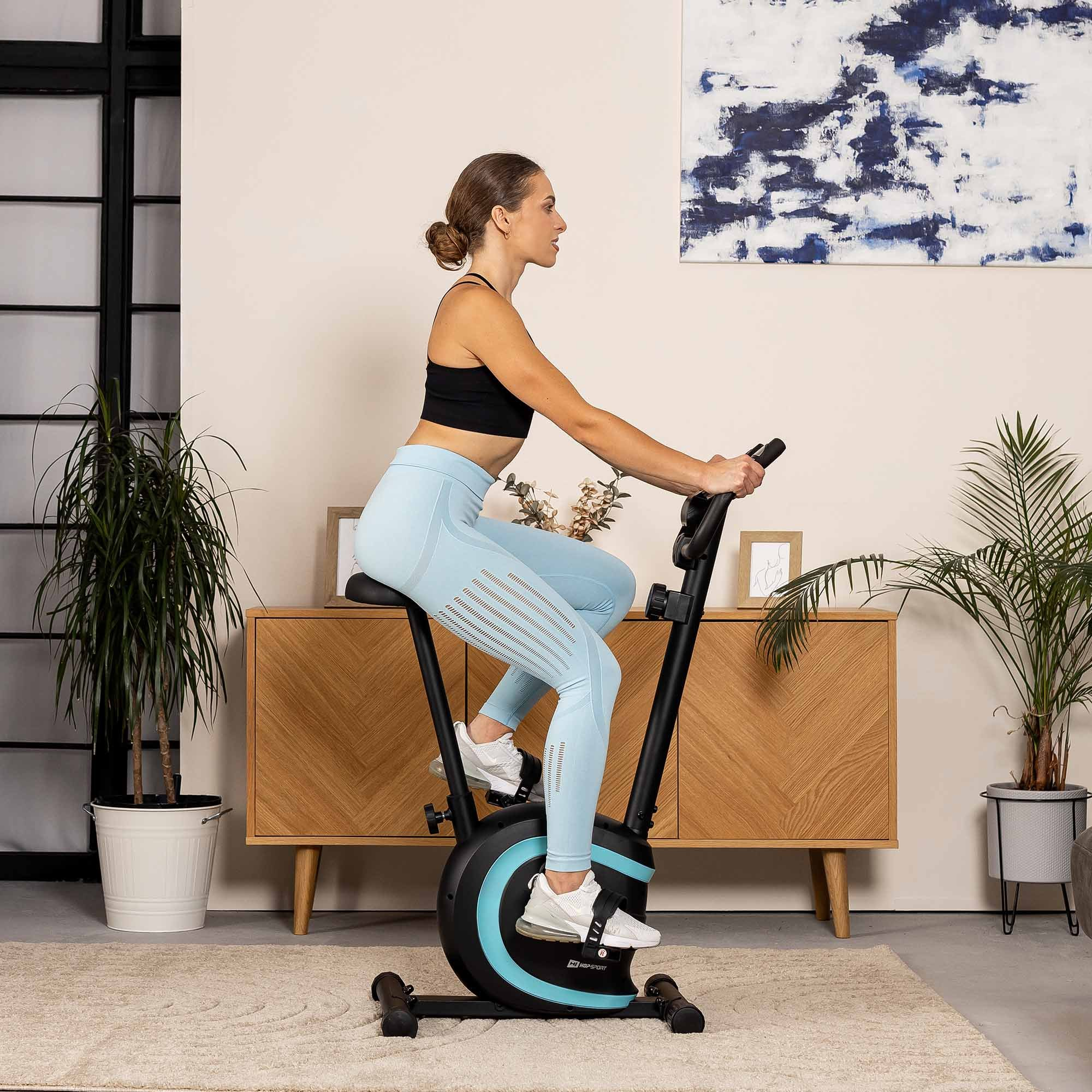 Eine blonde Frau in Sportkleidung trainiert auf einem Heimtrainer in einem modernen Zimmer mit Pflanzen.