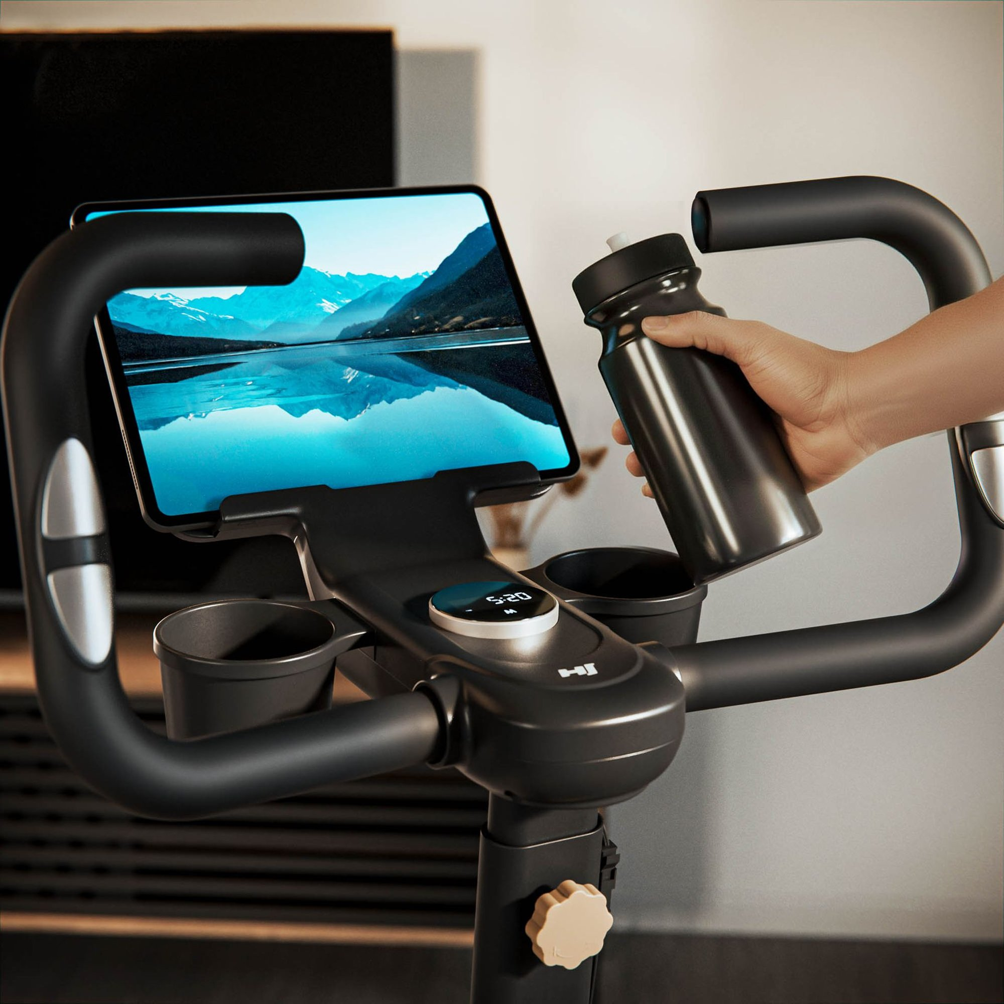 Eine Person hält eine Trinkflasche neben den Lenker eines Heimtrainers mit einem Tablet, das eine Berglandschaft zeigt.