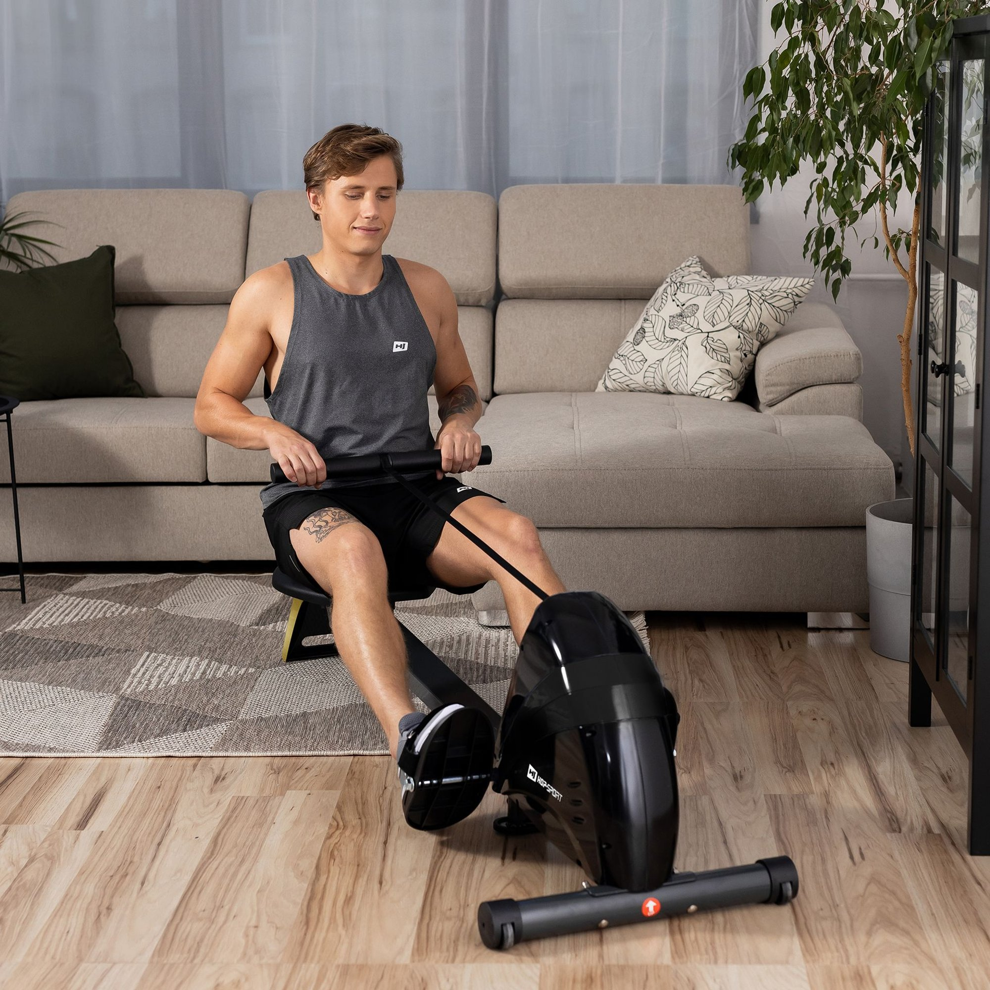 Ein Mann im grauen Tanktop trainiert auf einem schwarzen Rudergerät im Wohnzimmer.