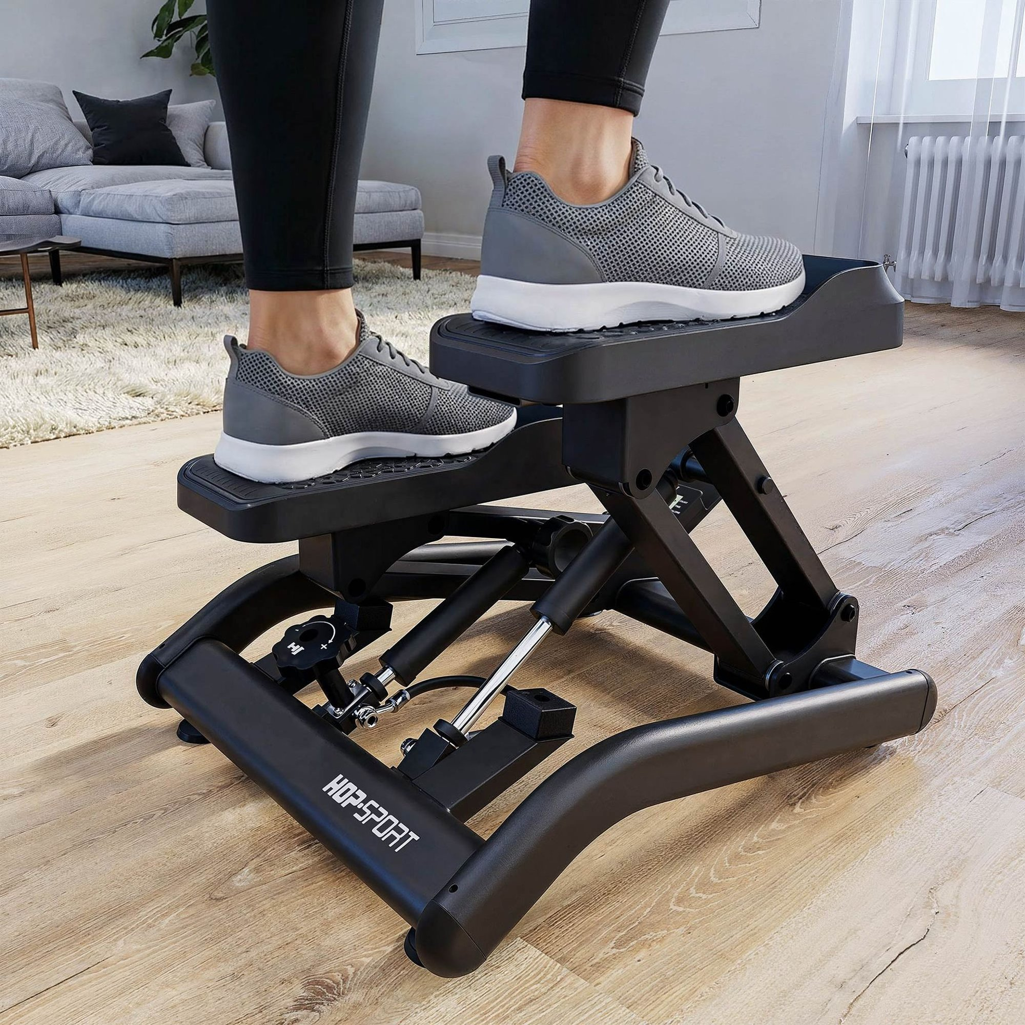 Person in schwarzen Leggings und grauen Sneakers nutzt einen schwarzen Heimtrainer-Stepper auf einem Holzboden.