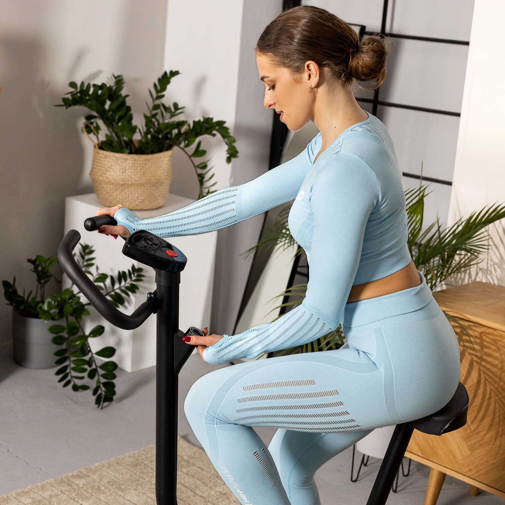 Eine Frau in hellblauer Sportkleidung trainiert auf einem Heimtrainer in einem hellen Raum mit Topfpflanzen.
