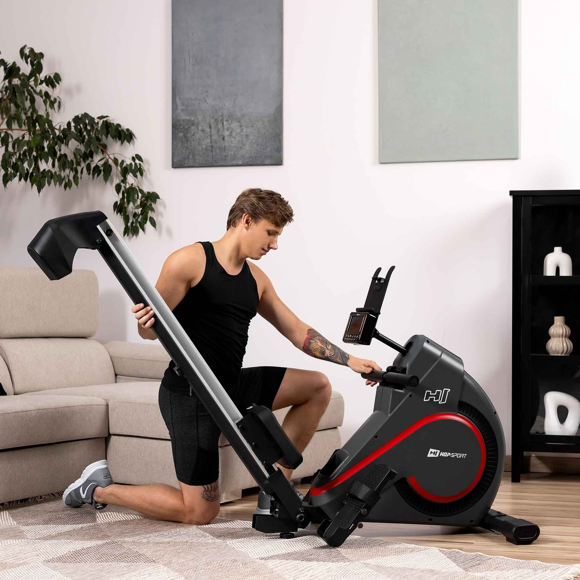 Ein Mann im Wohnzimmer klappt einen schwarzen Rudertrainer zusammen, im Hintergrund ein Sofa und Bilder.
