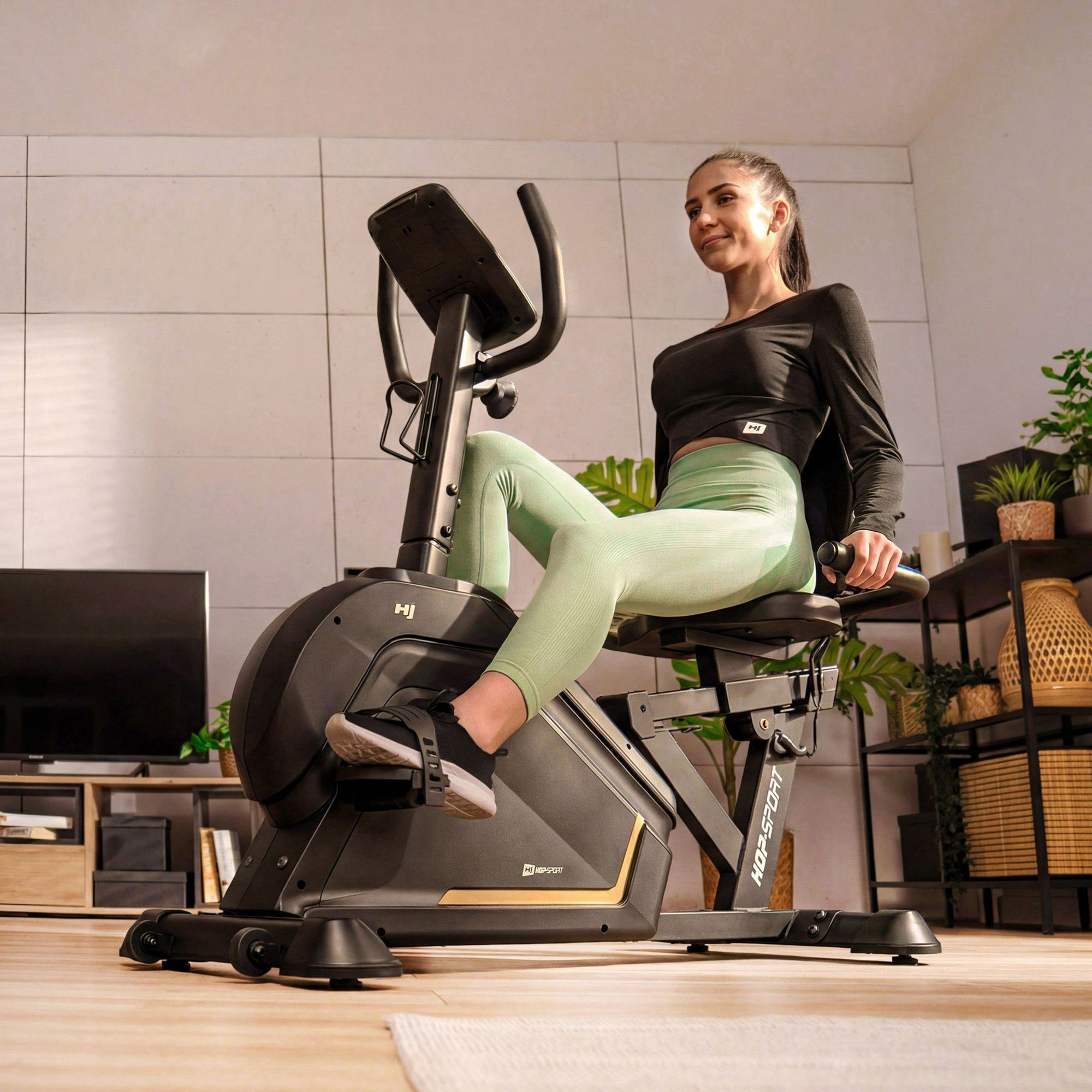 Eine Frau in Sportkleidung trainiert auf einem Liegeheimtrainer in einem modernen, pflanzengeschmückten Raum.
