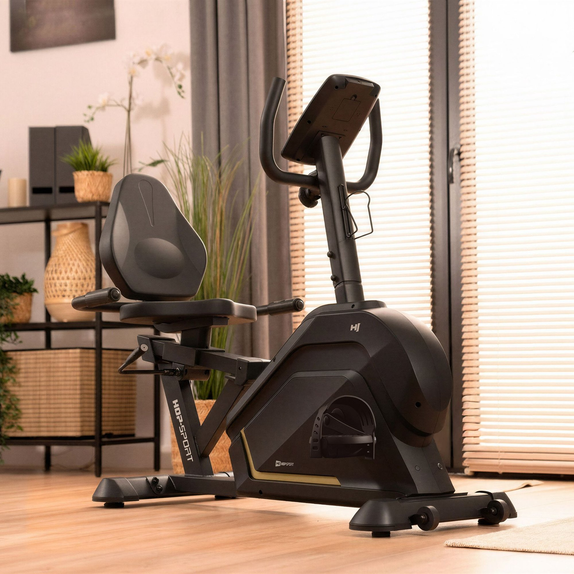 Ein schwarzer Liegeheimtrainer in einem hellen Wohnzimmer mit Regal und Pflanzen im Hintergrund.