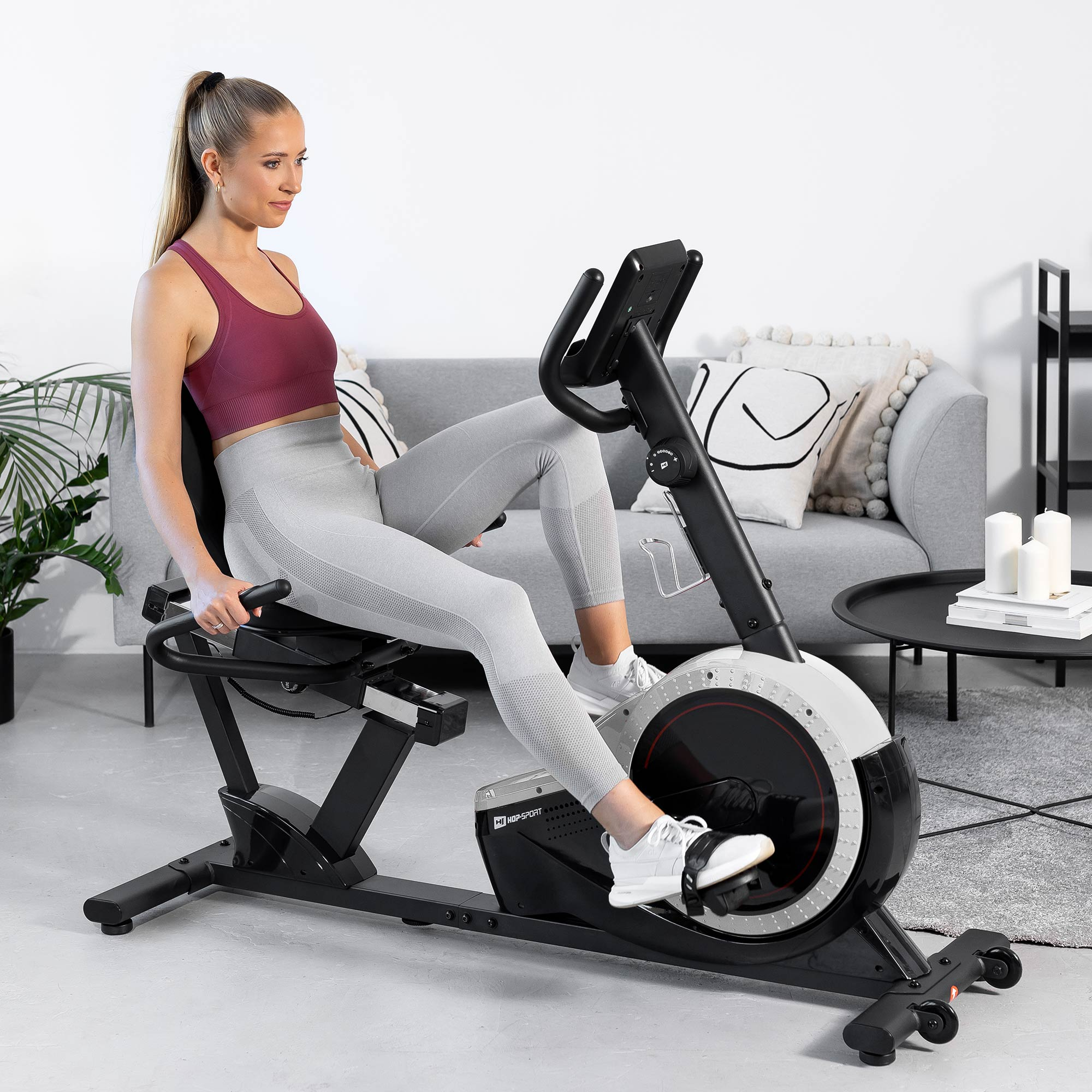 Eine blonde Frau in Sportkleidung sitzt auf einem Heimtrainer in einem hellen Raum mit Couch.