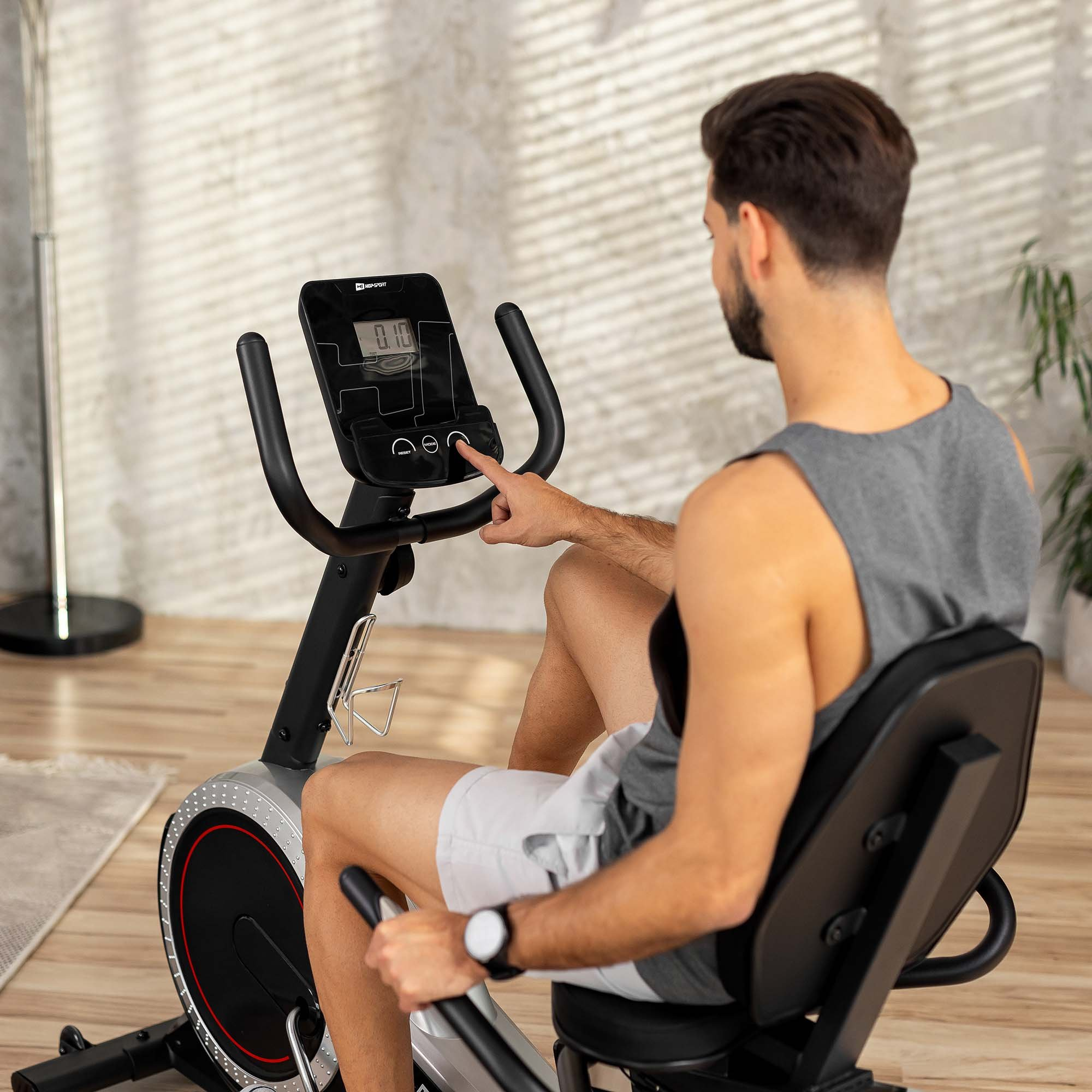 Ein Mann im grauen Tanktop sitzt auf einem Liegeheimtrainer und drückt das Tastenfeld.