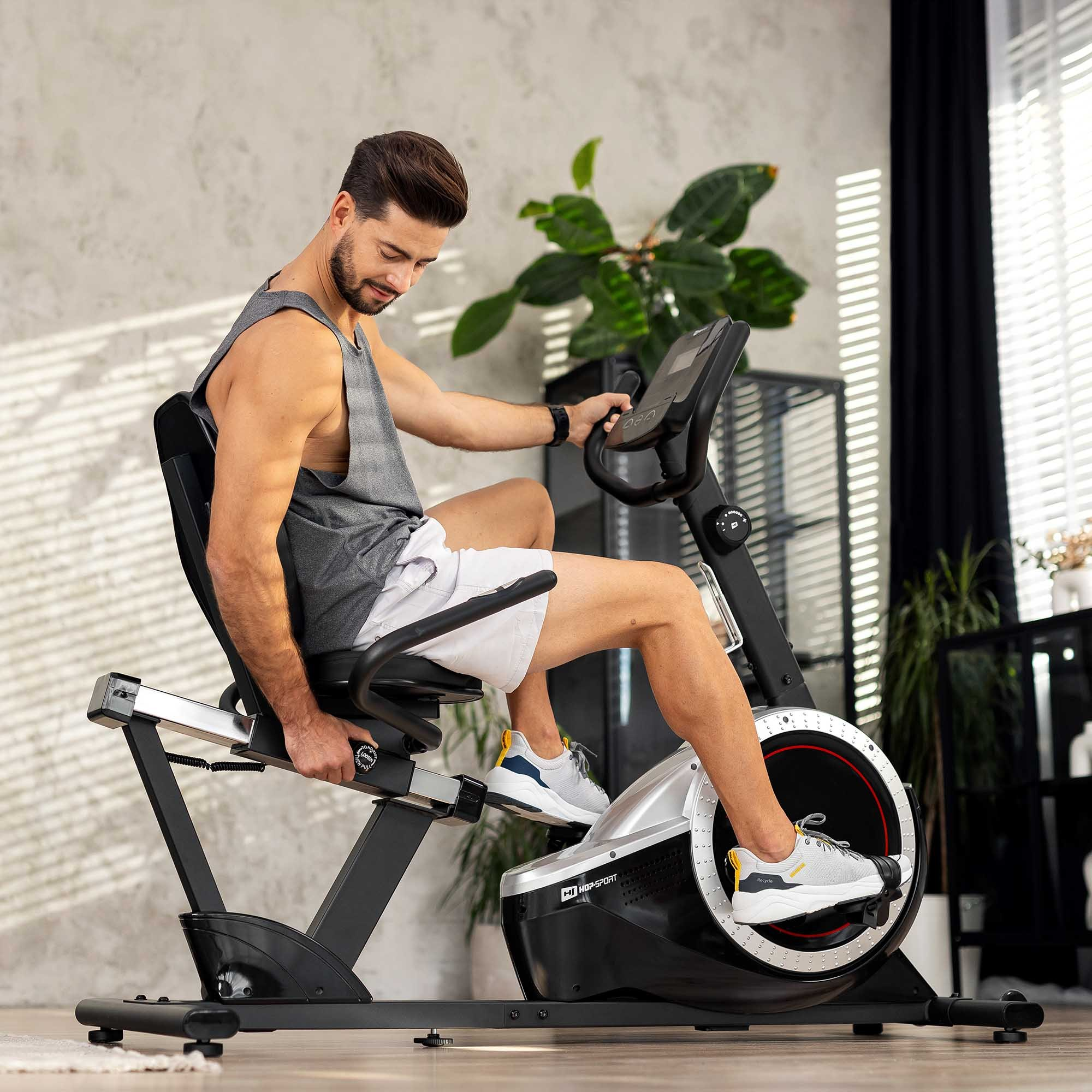 Ein Mann sitzt auf einem Heimtrainer in einem hellen Raum mit Pflanzen im Hintergrund und adjustiert den Sitz.