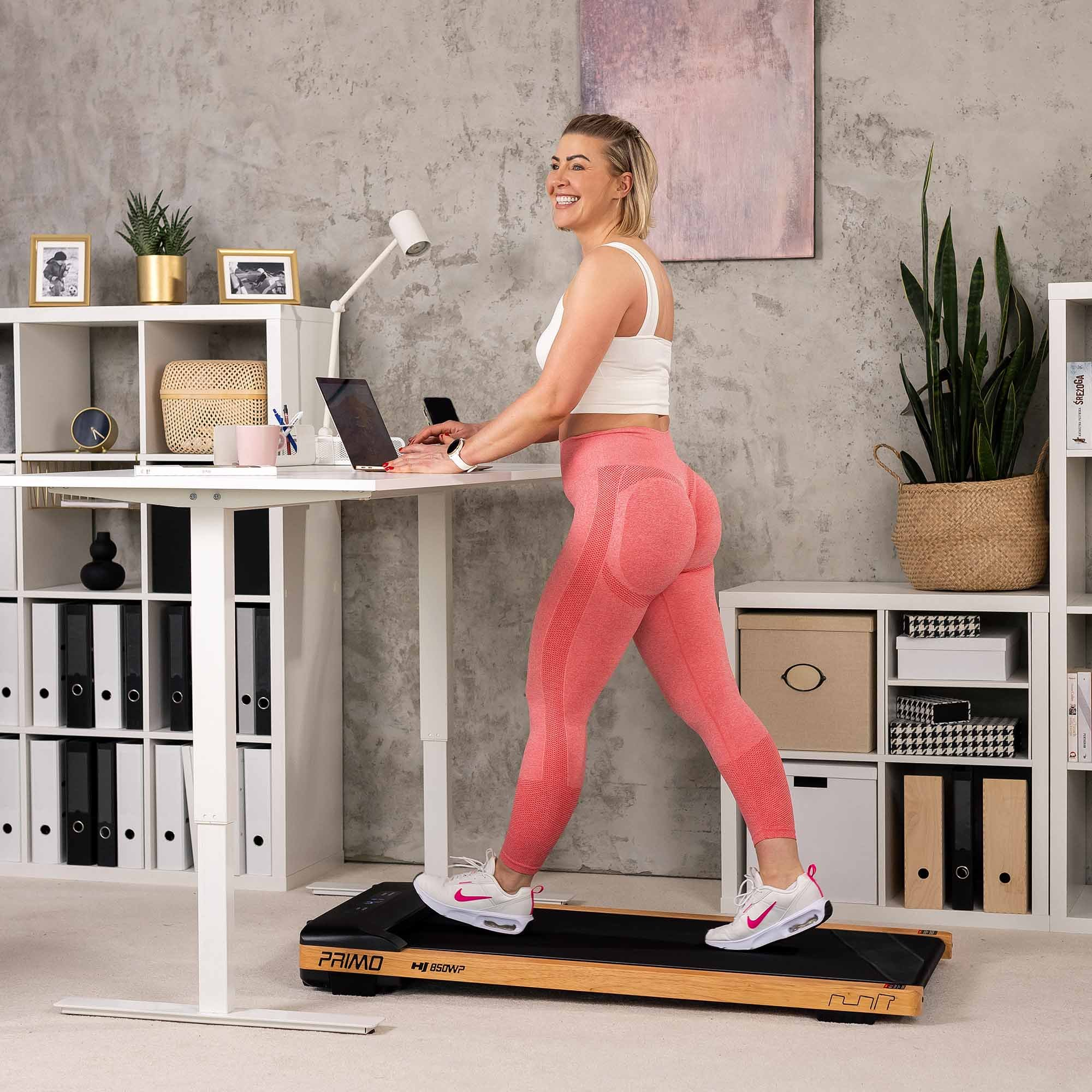 Eine lächelnde Frau in Sportkleidung geht auf einem Laufband, während sie an einem Stehpult arbeitet.