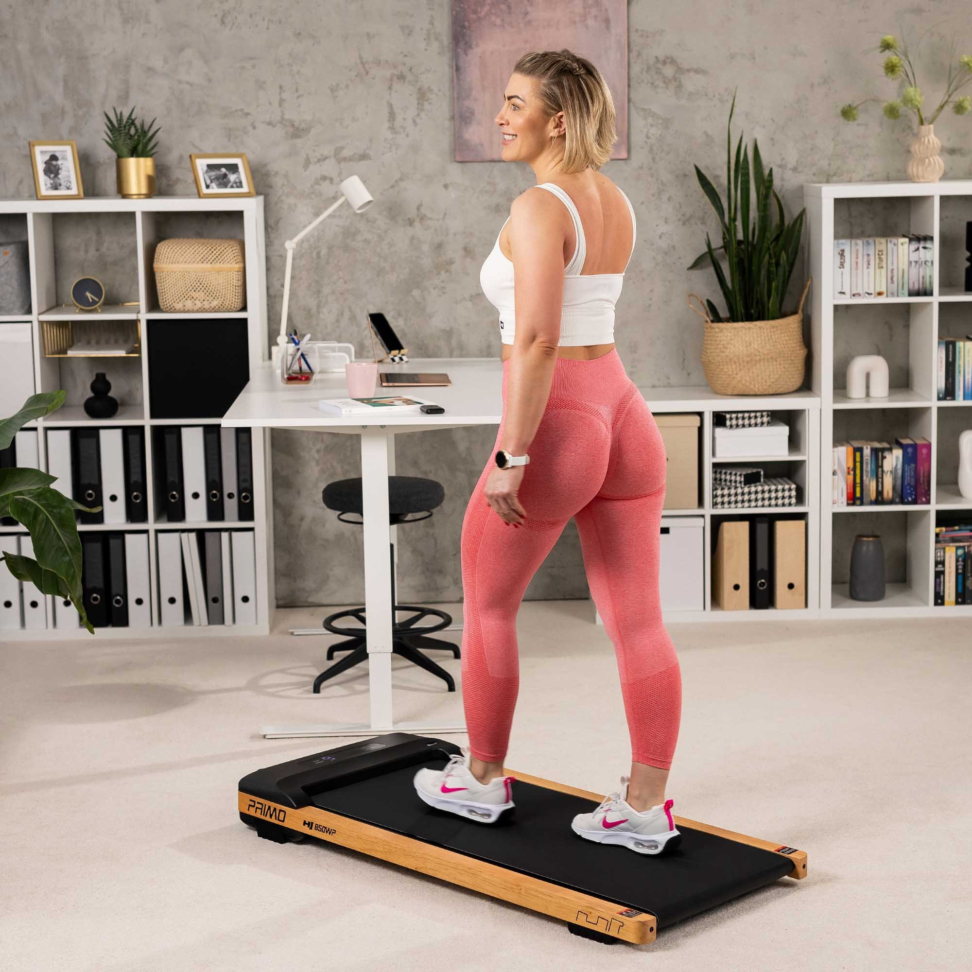 Eine blonde Frau im weißen Top und roten Leggings geht auf einem Laufband vor einem hellen Schreibtisch.